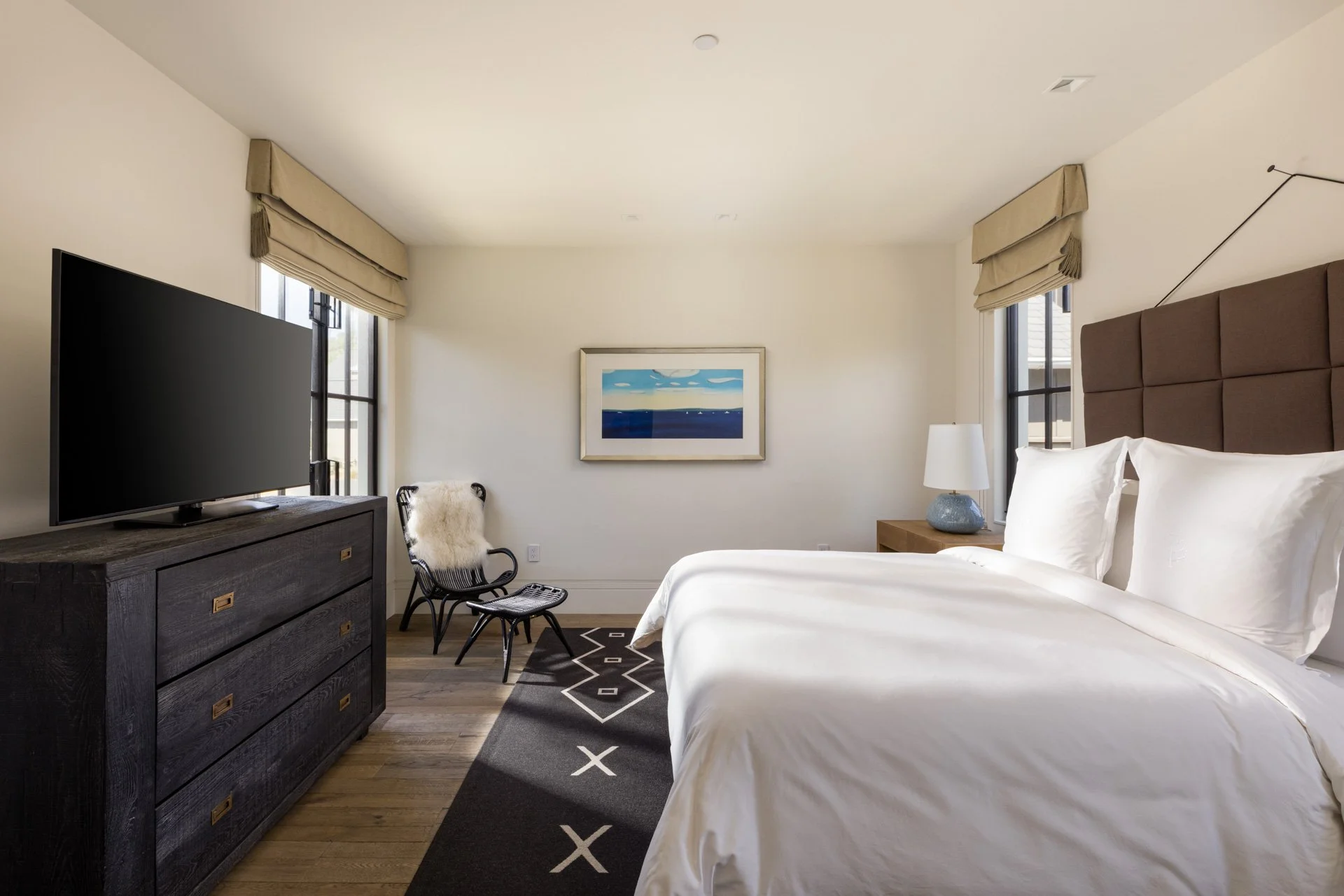Hotel room with a large bed, dark wooden dresser, flat-screen TV, chair with fur throw, window with shades, framed artwork, nightstand with lamp, and black rug with geometric pattern.