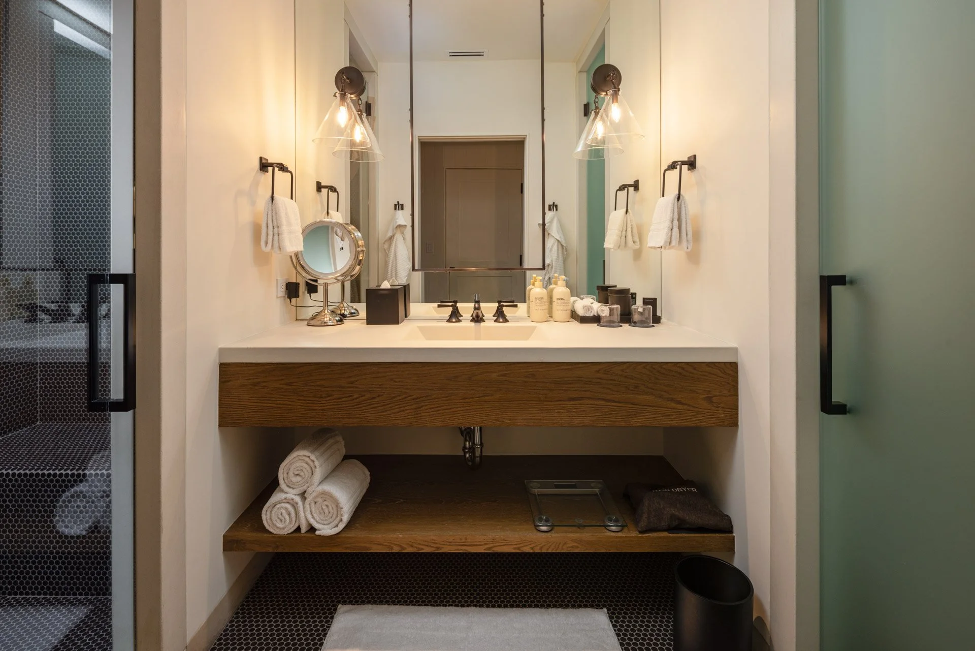 Modern bathroom vanity with a large mirror, two wall-mounted light fixtures, and a countertop with toiletries, a magnifying mirror, and neatly rolled towels underneath. There is a frosted glass door on the right and a glass shower door on the left.