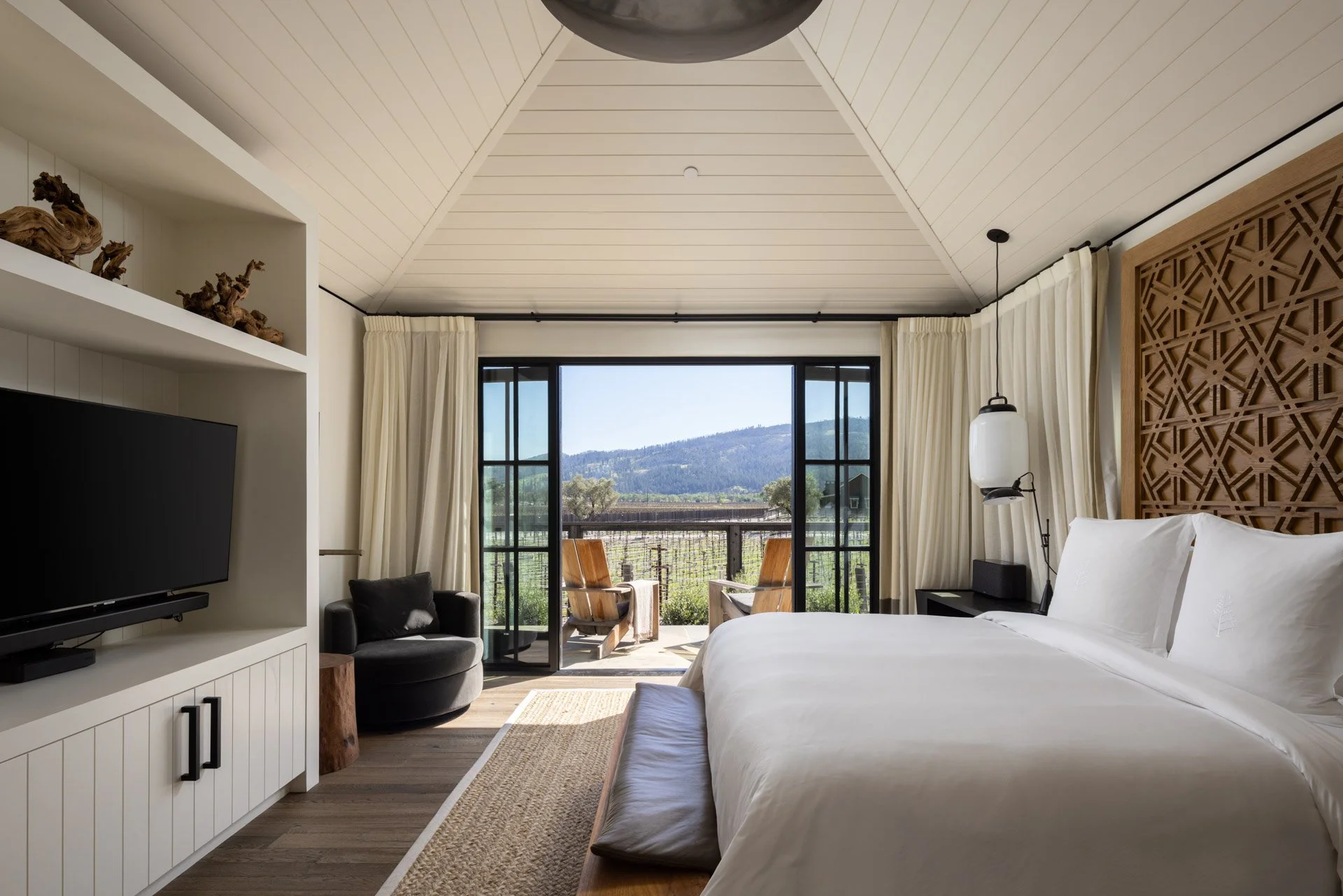 A modern bedroom with white bedding, a wooden headboard, and a balcony with outdoor chairs overlooking a scenic mountain landscape.