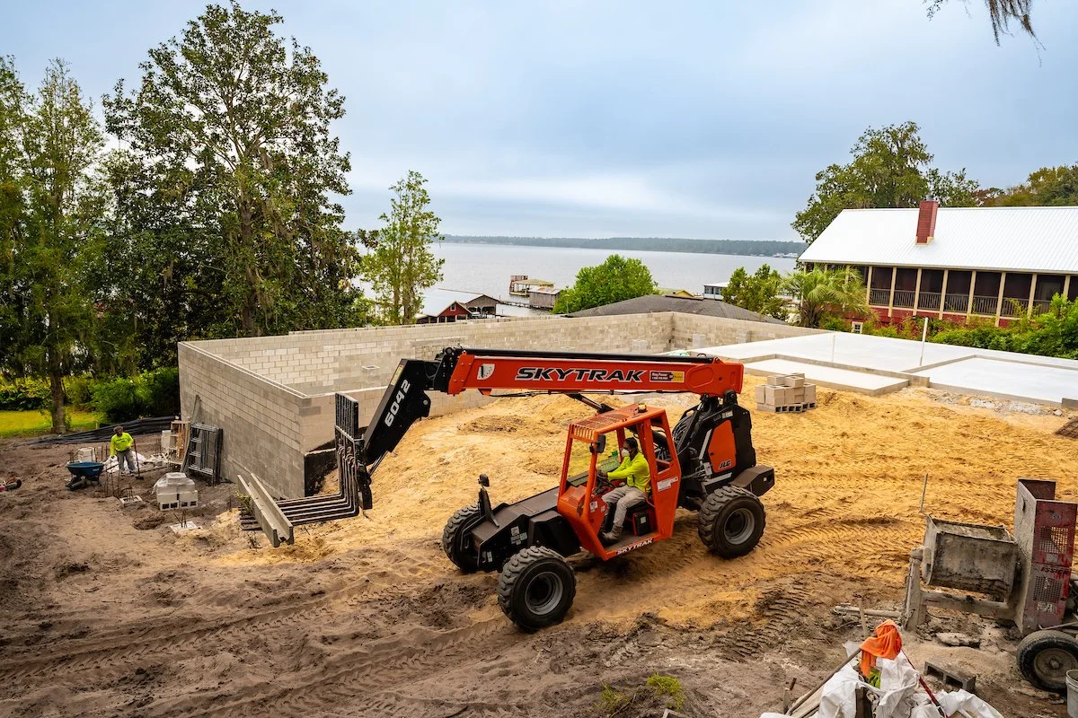 Construction site with a telescopic forklift moving soil, with workers and building materials around, overlooking a body of water and houses in the background.