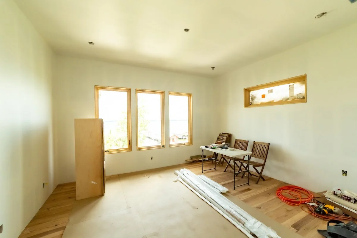 Room under construction with three windows, a small window higher on the wall, a table with tools, empty chairs, construction materials, and cables on the floor.