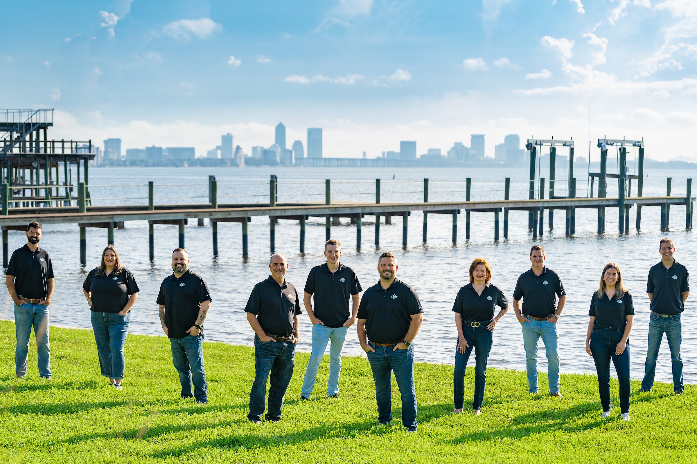 Group of ten people standing on a grassy area near a body of water with a city skyline and pier in the background.