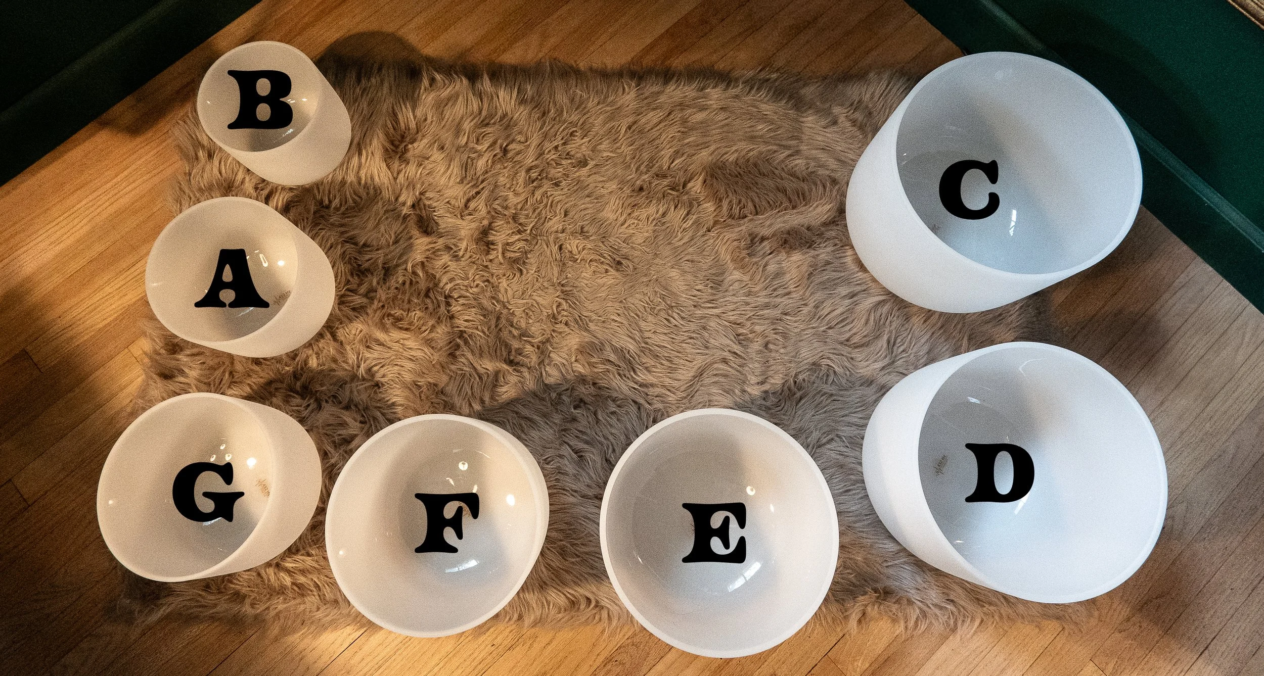 Seven crystal singing bowls labeled C, D, E, F, G, A and B arranged on a soft rug, showing the musical notes used during a sound bath.