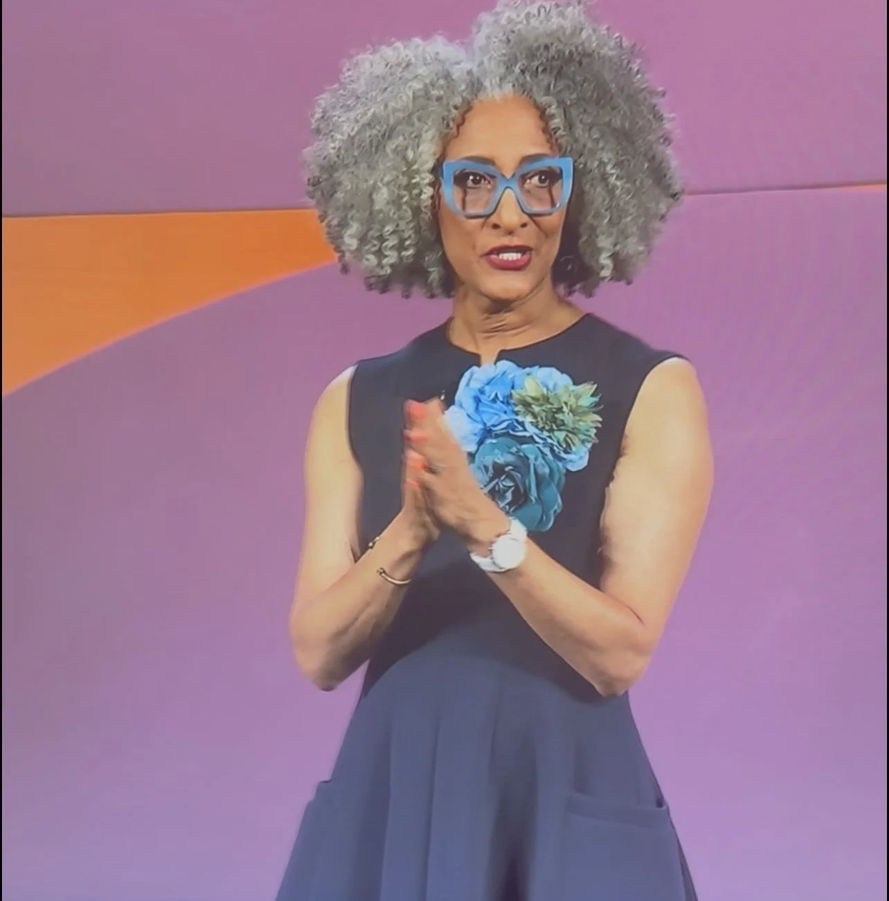 A woman with curly gray hair and blue glasses standing with her hands pressed together, wearing a sleeveless black dress with a blue floral design in front, against a purple and orange background.