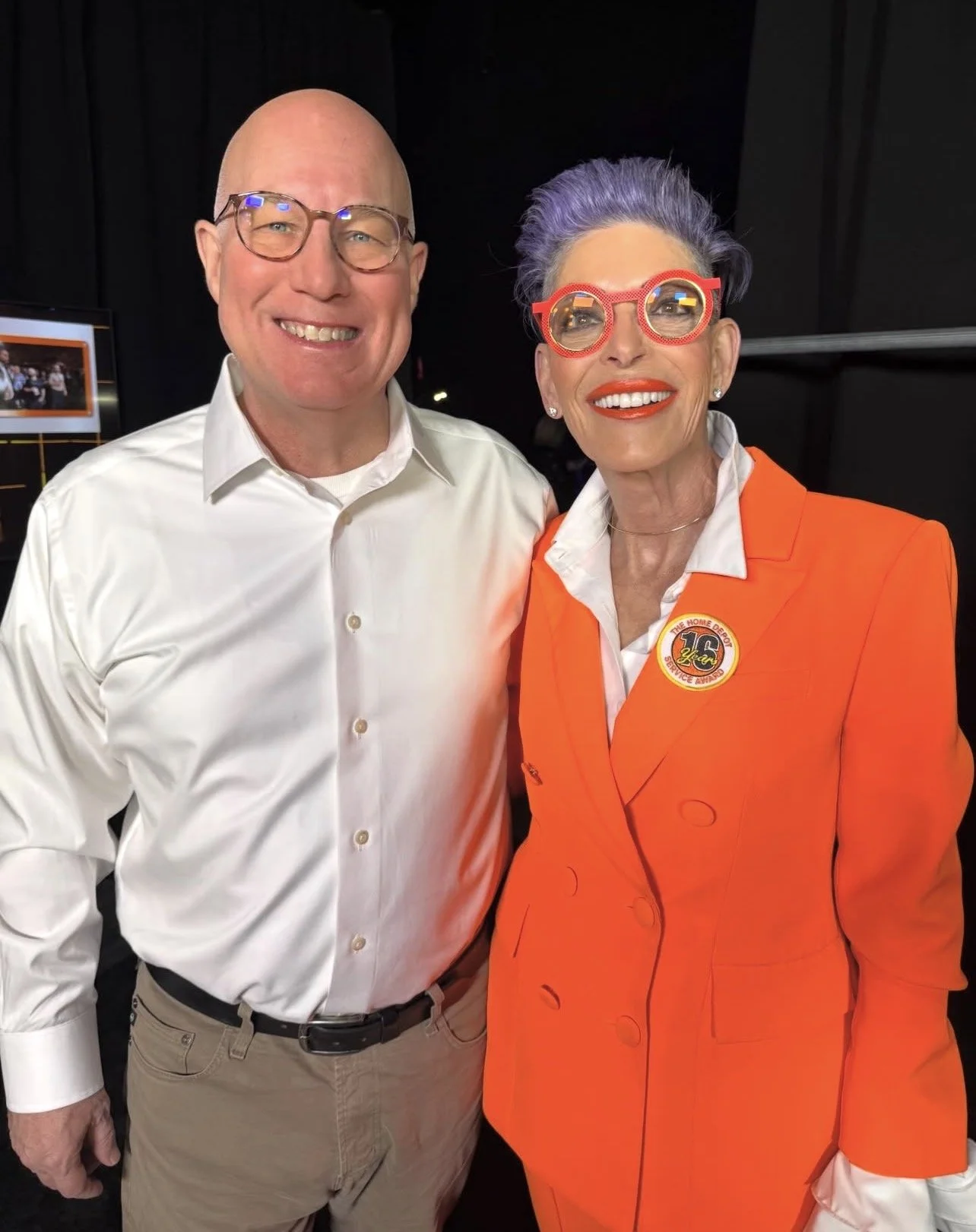 A man and a woman smiling at the camera, dressed in colorful attire. The woman is wearing a bright orange blazer with a badge and pink glasses, and has purple hair. The man is dressed in a white shirt and beige pants, with glasses, and is bald.