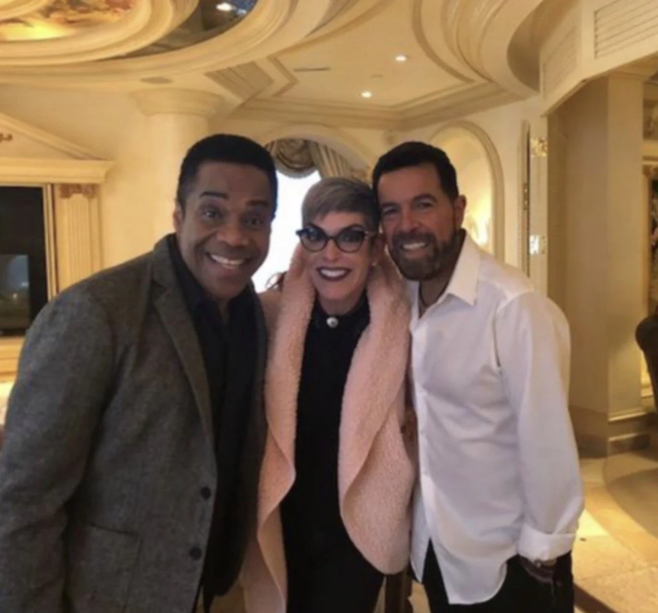 Three people smiling and posing together indoors, with elegant decor and chandelier lighting in the background.