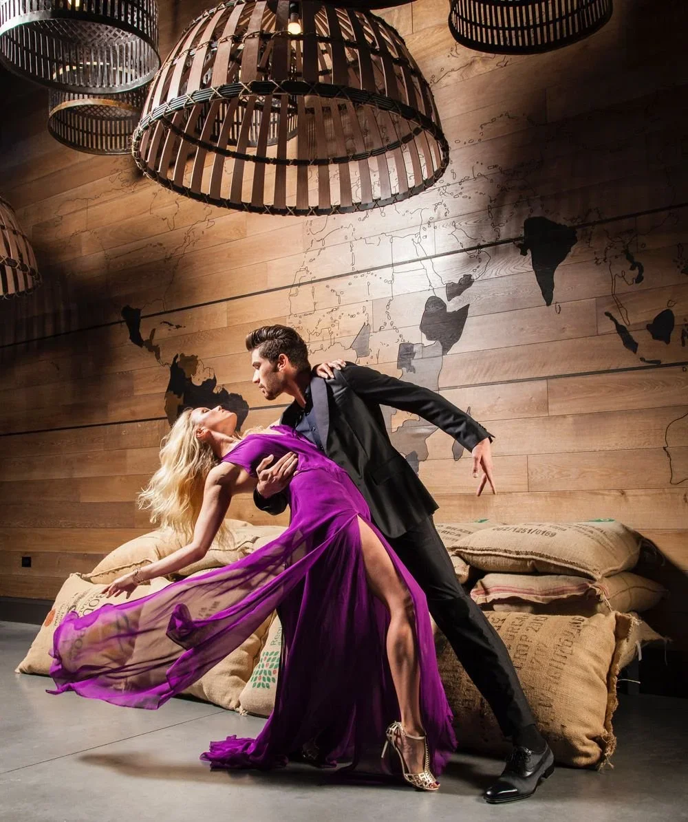 A man and woman dance passionately in a stylish, dimly lit room with wooden walls and a world map decoration. The woman wears a flowing purple dress with a high slit and gold high heels. The man is dressed in a black suit. Background features large h