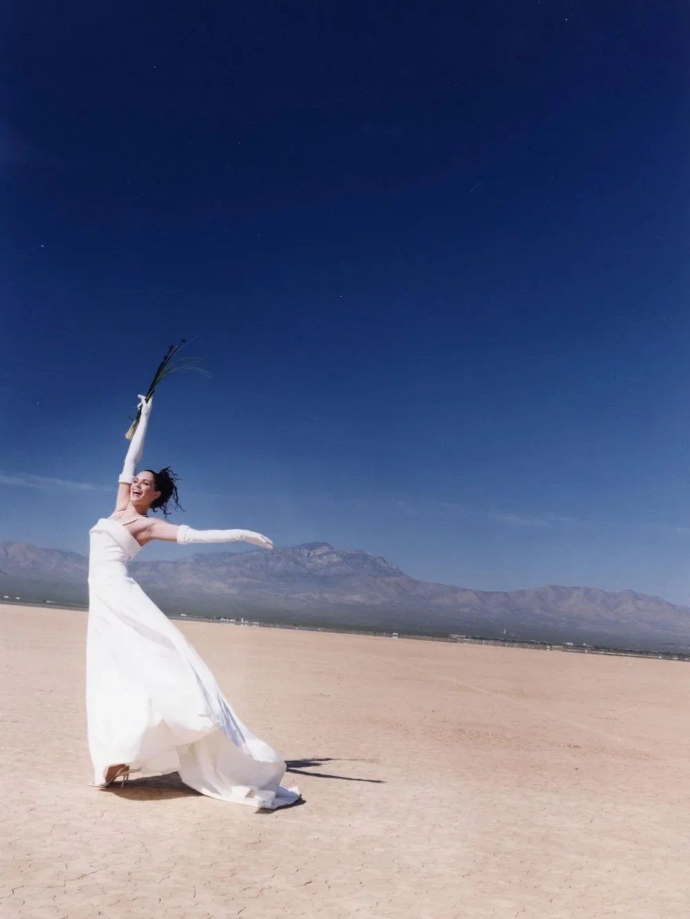 Woman in a white dress and long gloves dancing or celebrating in a desert landscape under a clear blue sky, mountains in the background, holding a bouquet.