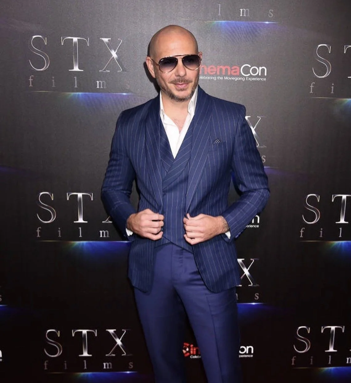 A man in a blue pinstripe suit, white shirt, and sunglasses posing in front of a black backdrop with 'STX films' and 'Cinemacon' logos.