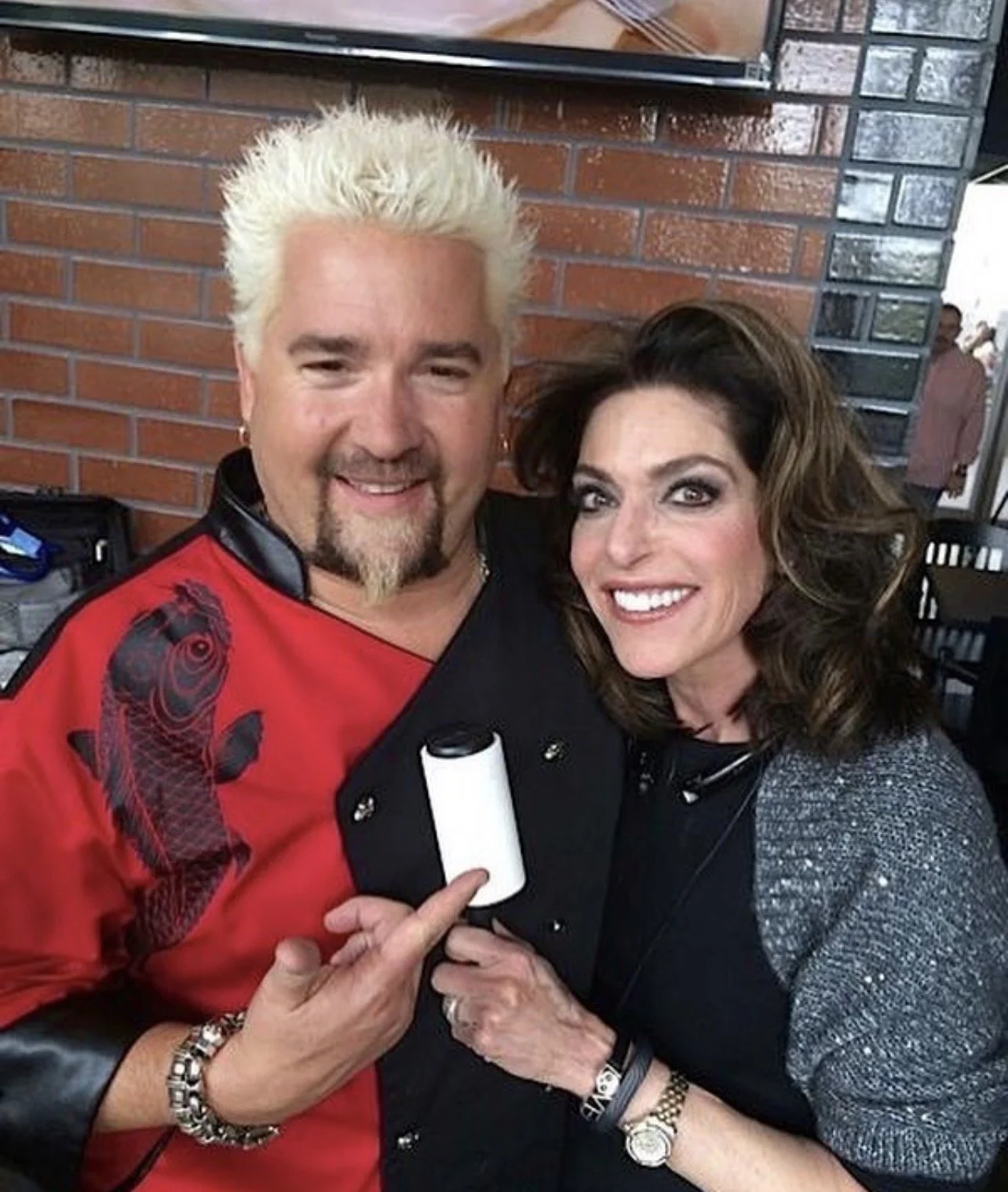 A man with spiked bleached blonde hair and a goatee, and a woman with wavy brown hair, smiling, holding a small white and black cylindrical object, standing in front of a brick wall.