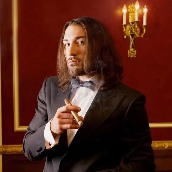 A man with long hair and a beard dressed in a black tuxedo and bow tie, holding a cigar, posing in front of a red wall with a gold frame and a lit candelabra in the background.