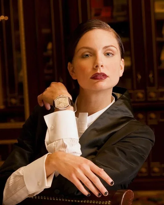 A woman dressed as a lawyer or judge, with dark lipstick and short dark hair, sitting at a desk with a confident expression, adjusting her suit jacket in a wood-paneled office.