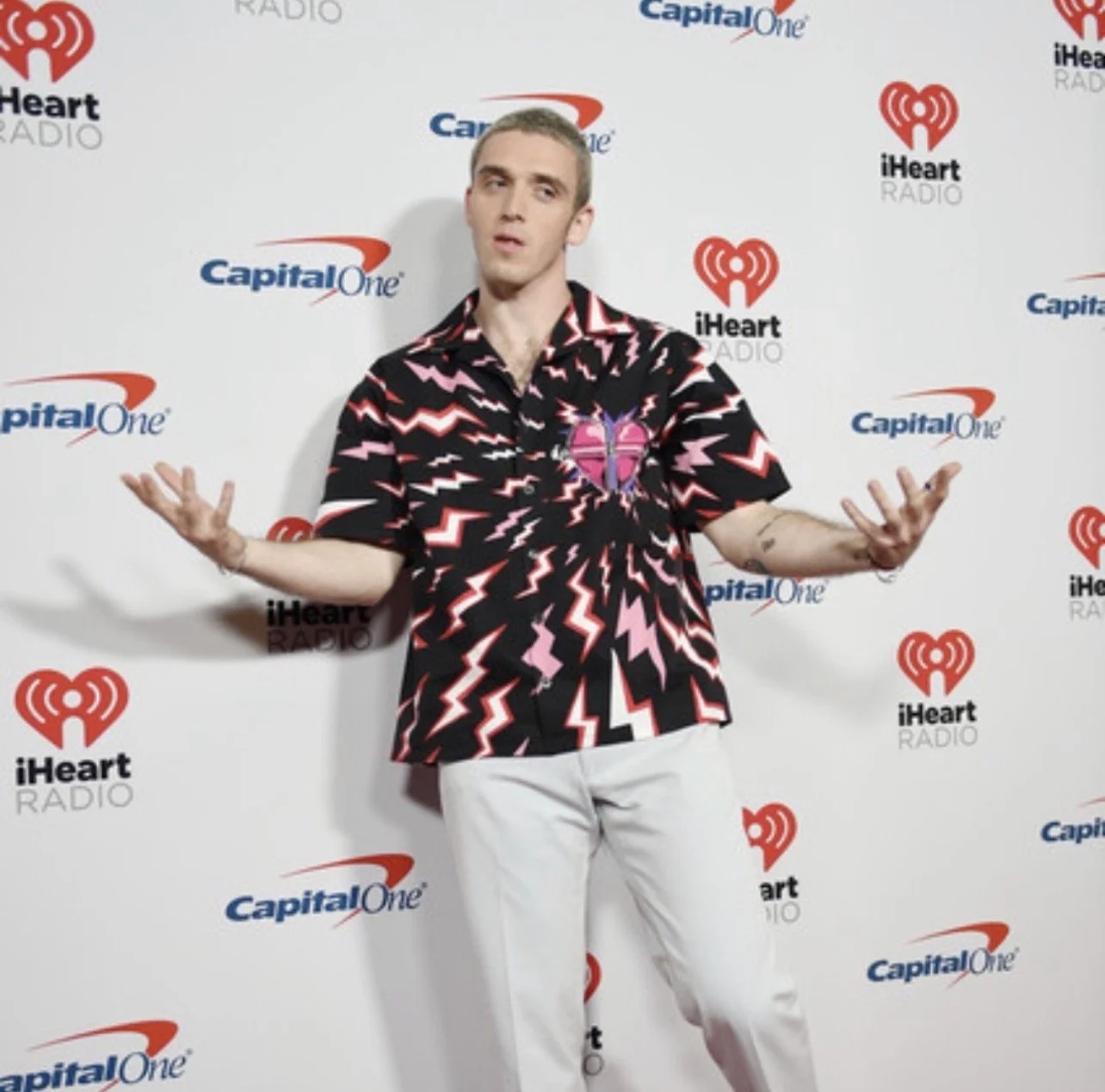 A man standing in front of a backdrop with iHeartRadio and Capital One logos, wearing a black shirt with a pink, red, and white lightning pattern and a broken heart graphic, gesturing with his hands.