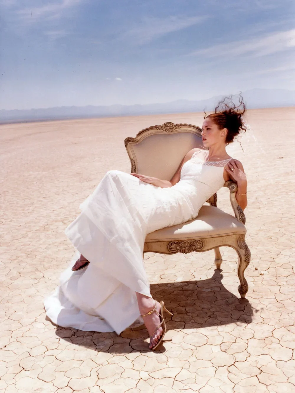 Woman in a white dress lounging on an ornate vintage sofa in a dry, cracked desert landscape under a blue sky.