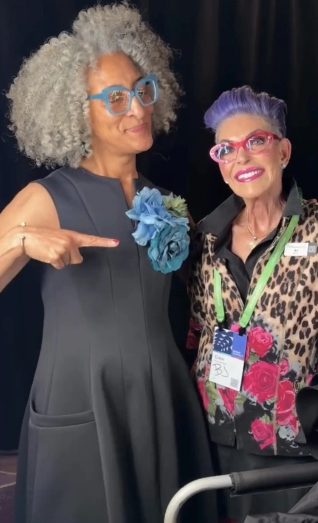 Two women with colorful glasses and vibrant outfits standing together against a dark background. One wears a black dress with blue flowers, the other has a leopard print jacket with pink roses and a name tag.