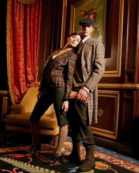 A woman leans her head on a tall man in a room with red curtains, a painting, and ornate wood paneling.