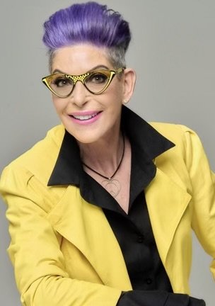 Woman with purple hair, wearing cat-eye glasses, a black shirt with a yellow collar, and a yellow blazer.