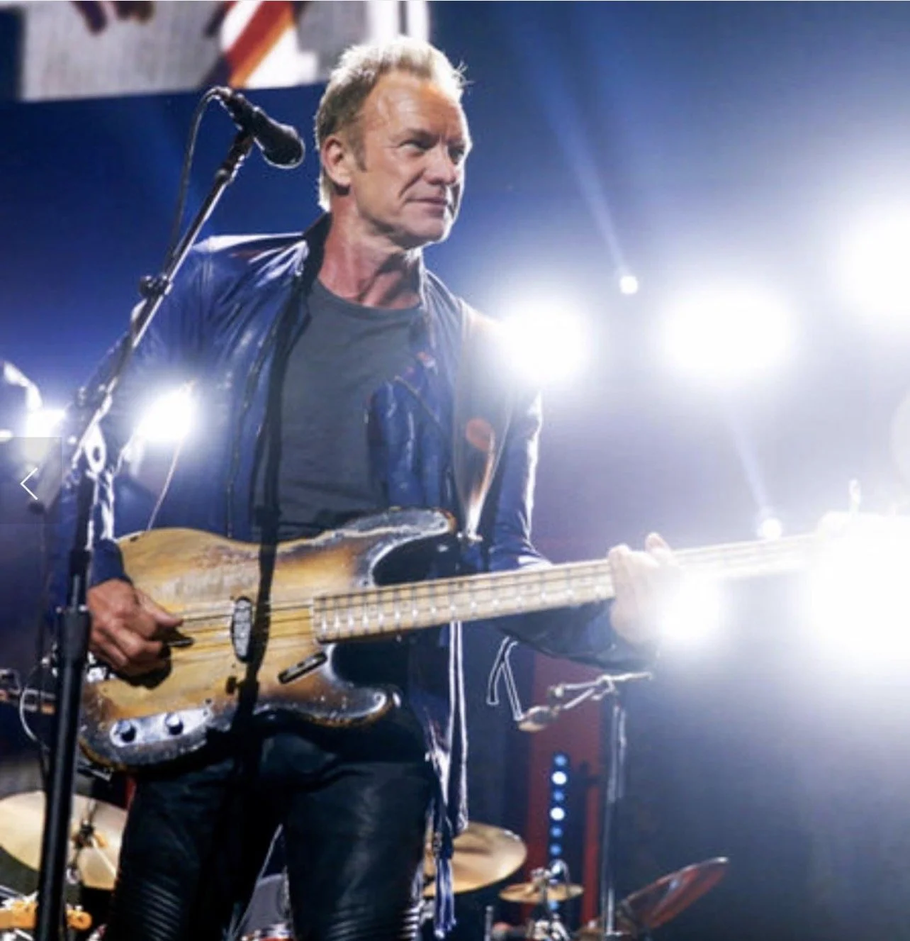 A man playing electric guitar on stage, wearing a leather jacket, with bright stage lights behind him.