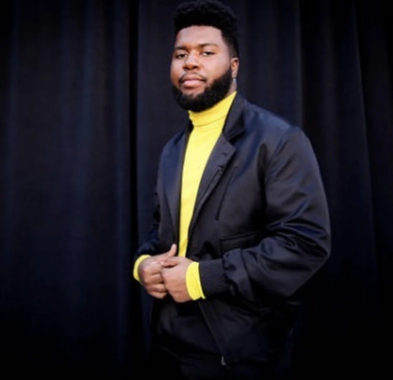 A man with a beard and short hair standing against a black curtain background, wearing a black jacket over a yellow turtleneck sweater.