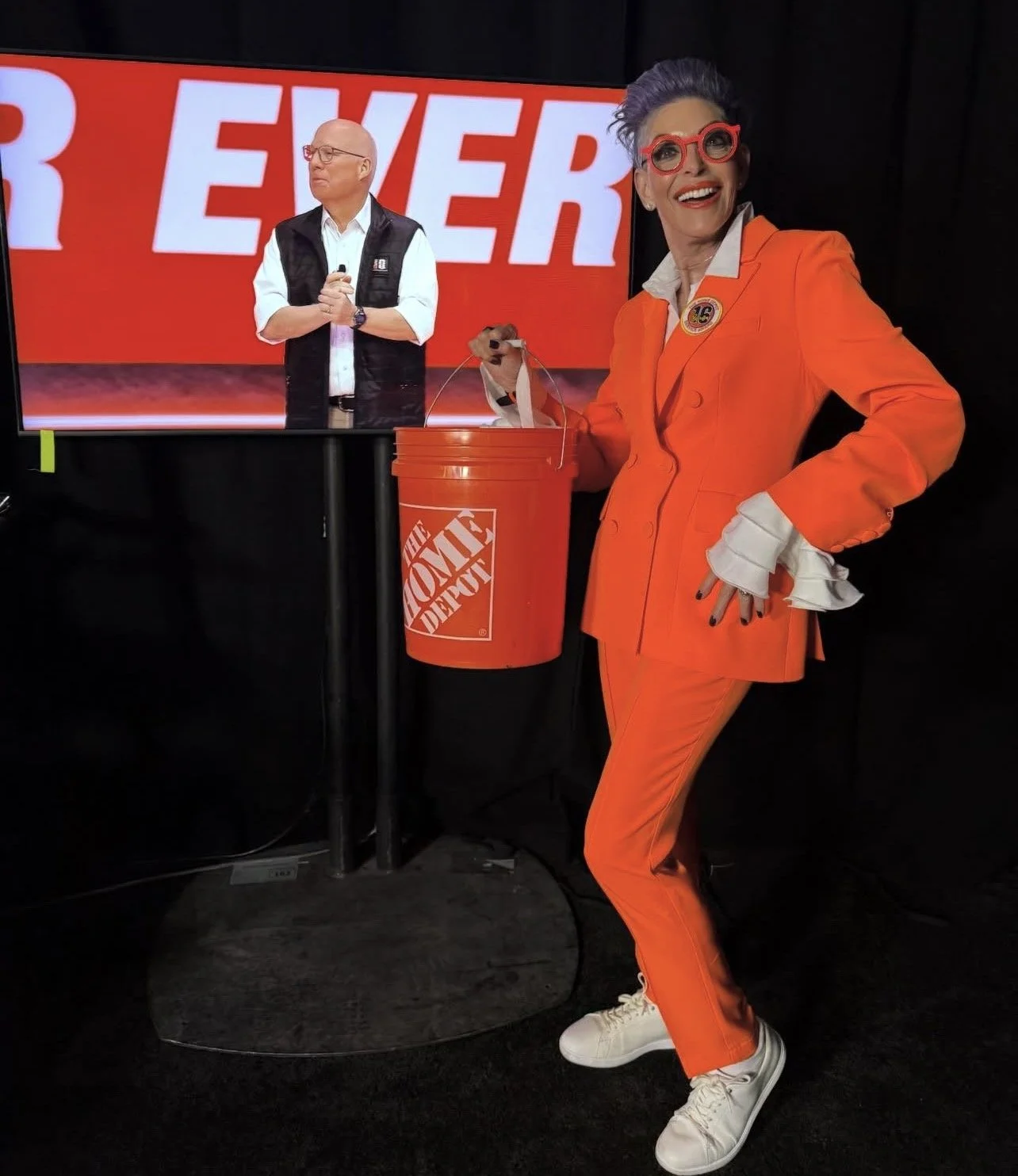 A woman dressed in an orange suit with white cuffs and white sneakers is smiling and holding a Large orange Home Depot bucket with a stuffed animal inside. In the background, a man is on a screen, wearing glasses and a black vest, with large white and red text behind him.