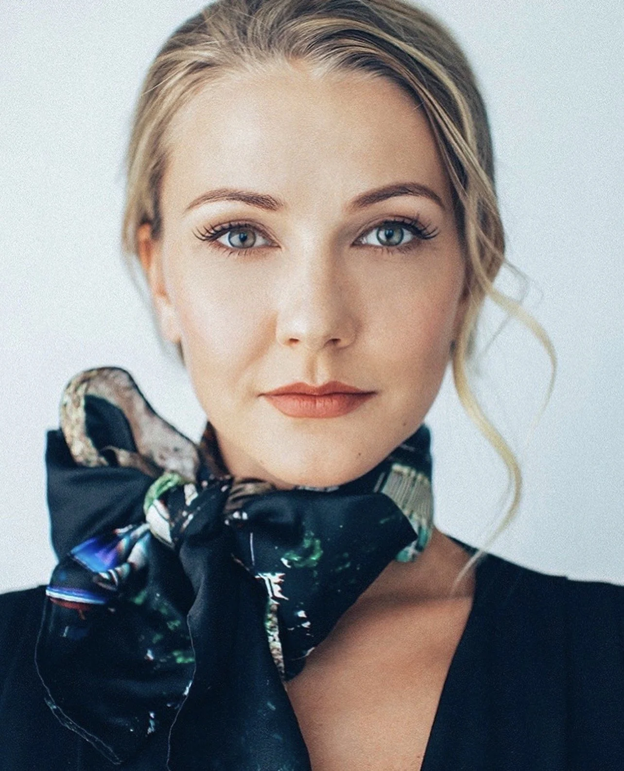 Close-up portrait of a woman with blonde hair, blue eyes, and makeup. She is wearing a black top and a colorful, patterned scarf tied around her neck.