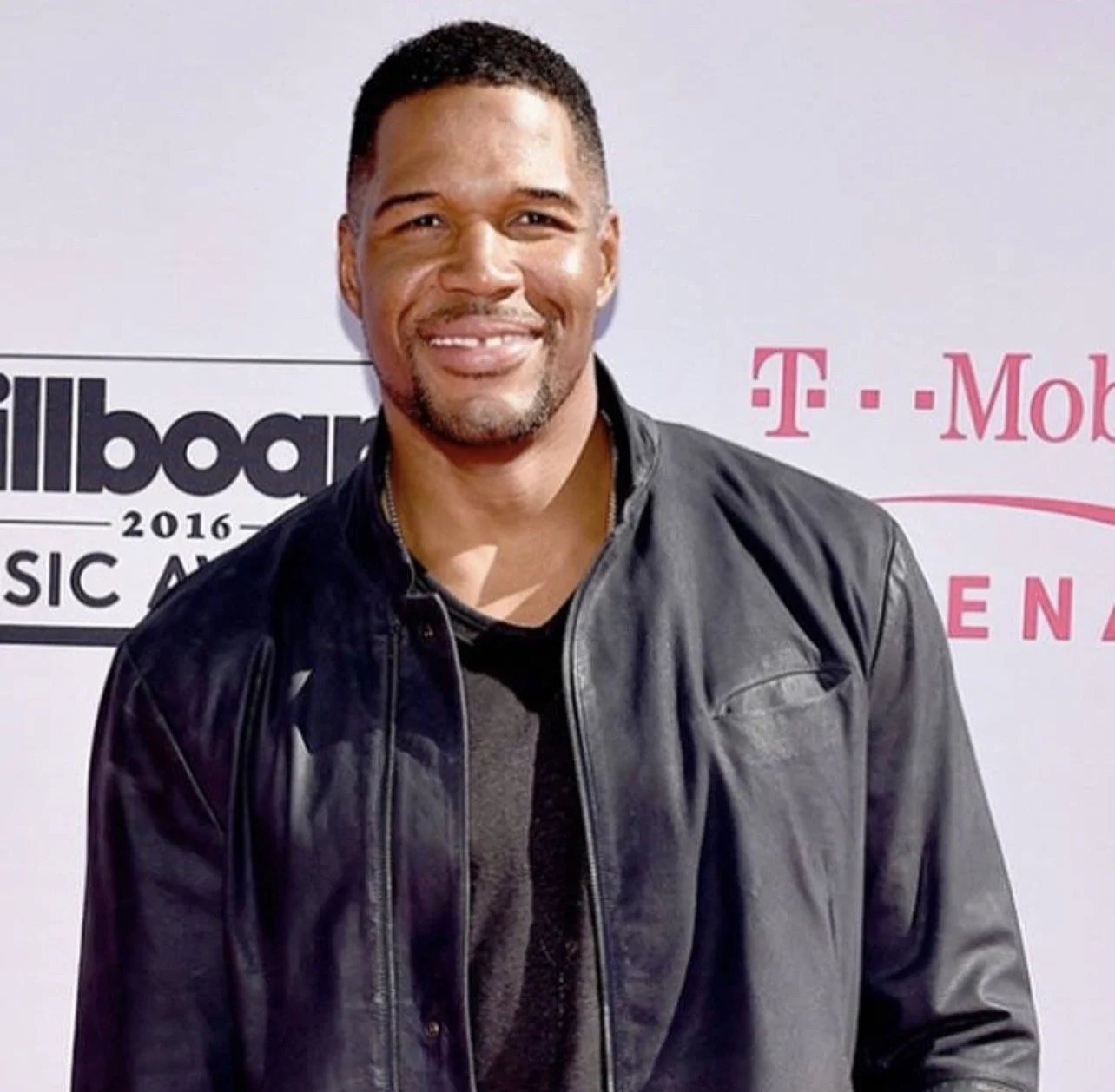 A man smiling at a music awards event, wearing a black leather jacket and black shirt, with logos for Billboard Music Awards 2016 and T-Mobile in the background.