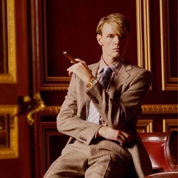 A young man with blond hair sitting in a luxurious room with dark wooden paneling, wearing a beige plaid suit, holding a cigar in his right hand while gazing at the camera.