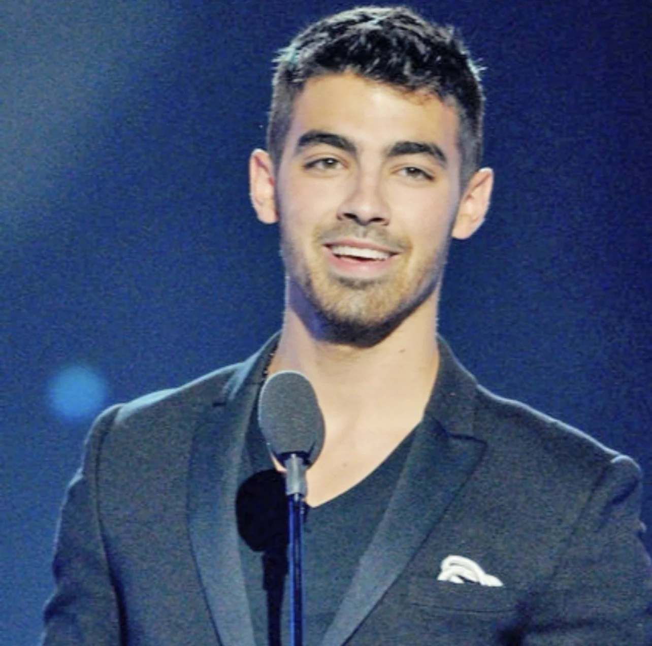 A young man with dark hair and a beard stands in front of a microphone on a stage, wearing a black suit jacket and a black shirt, smiling at the camera.