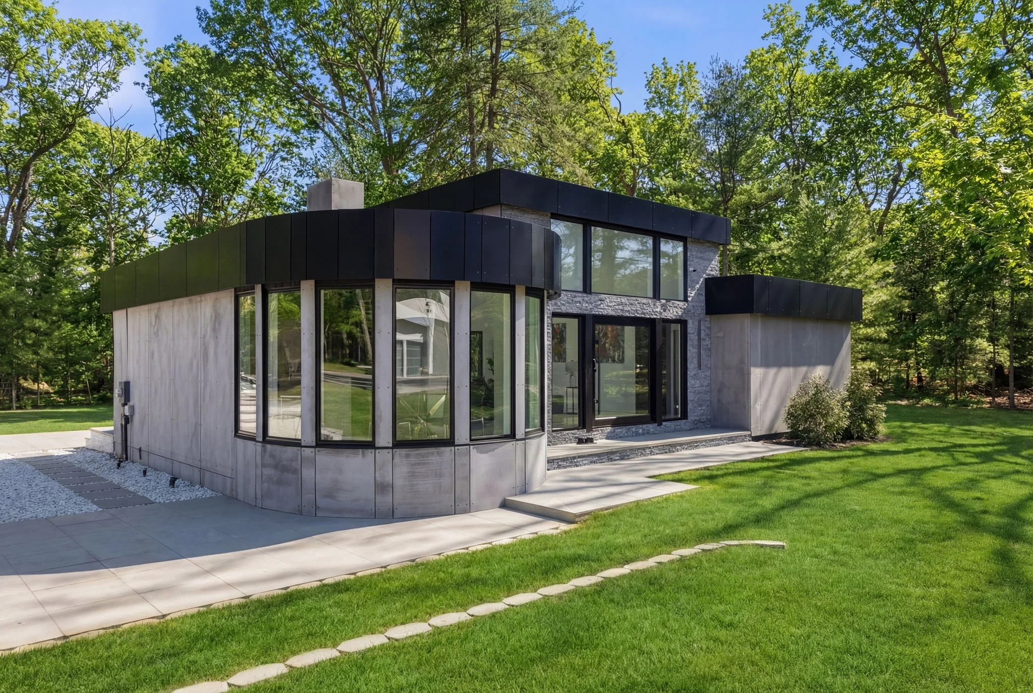 Modern house with curved concrete walls, large windows, and black metal accents, surrounded by green grass and trees under a clear blue sky.