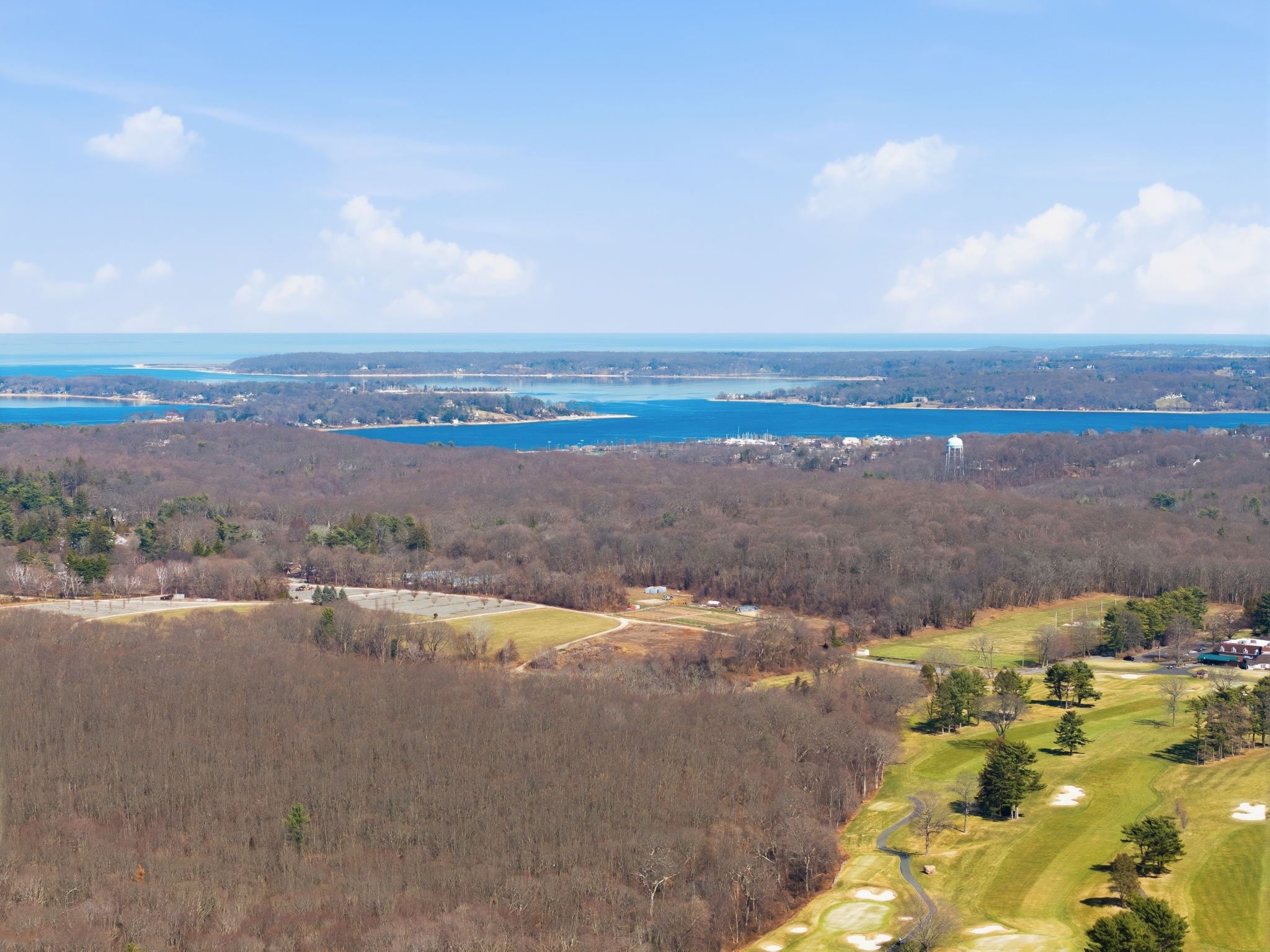 Aerial view of a landscape with a golf course, wooded areas, a lake, and blue sky with clouds.