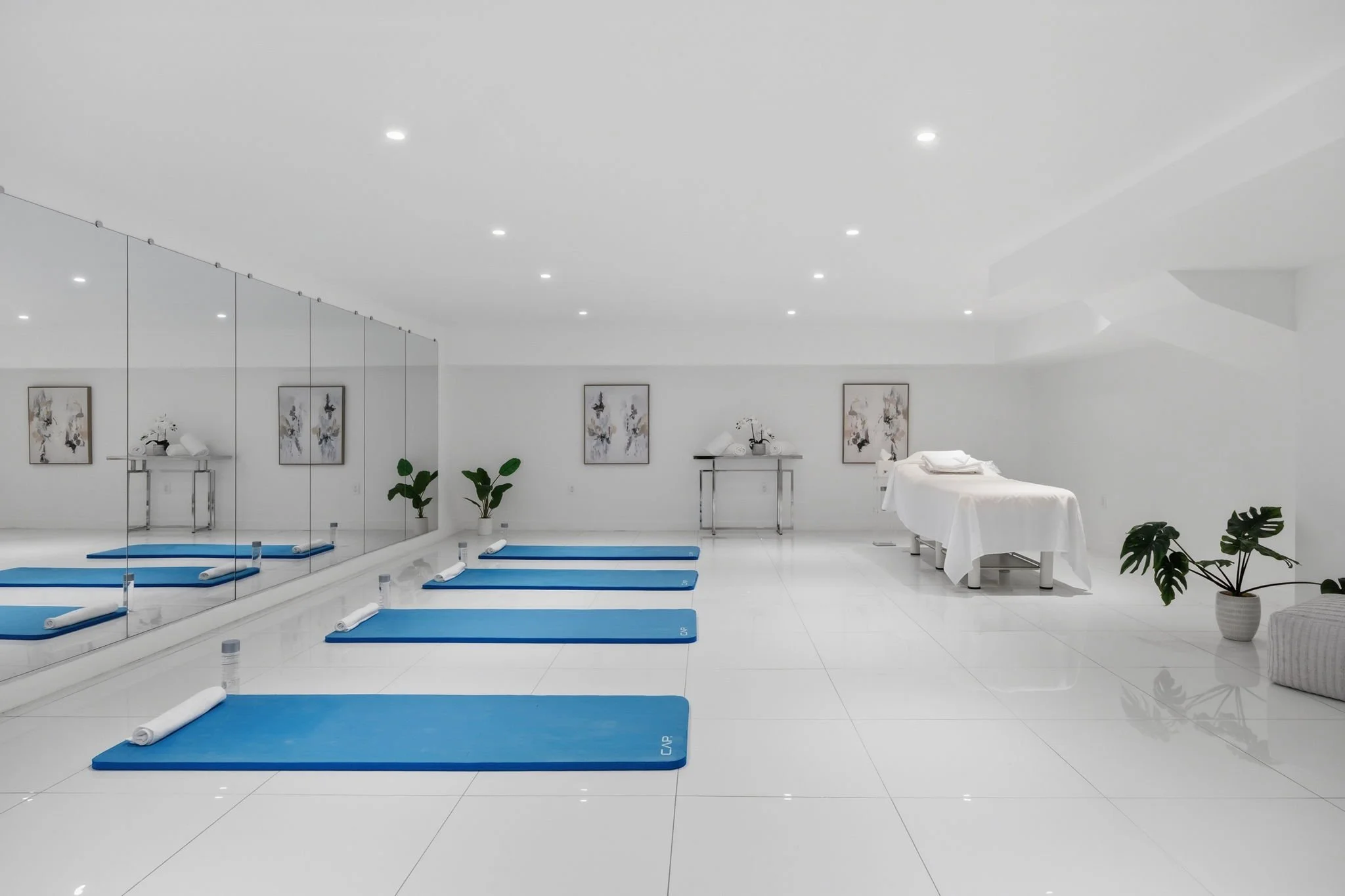 A spacious, minimalist wellness or yoga studio with white walls and floor, blue yoga mats arranged in a line, a large mirror on one wall, and potted plants for decor.