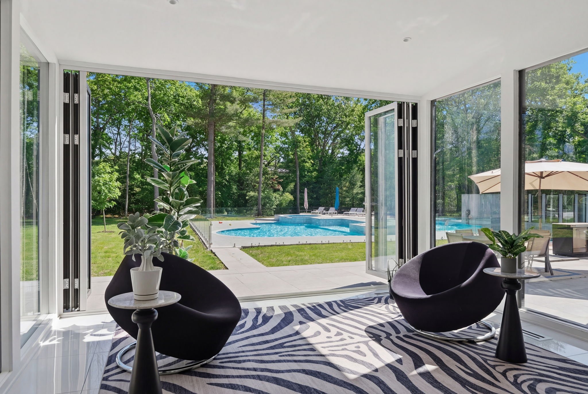 Living room with black modern chairs, plants, and a view of a backyard with a pool and trees.