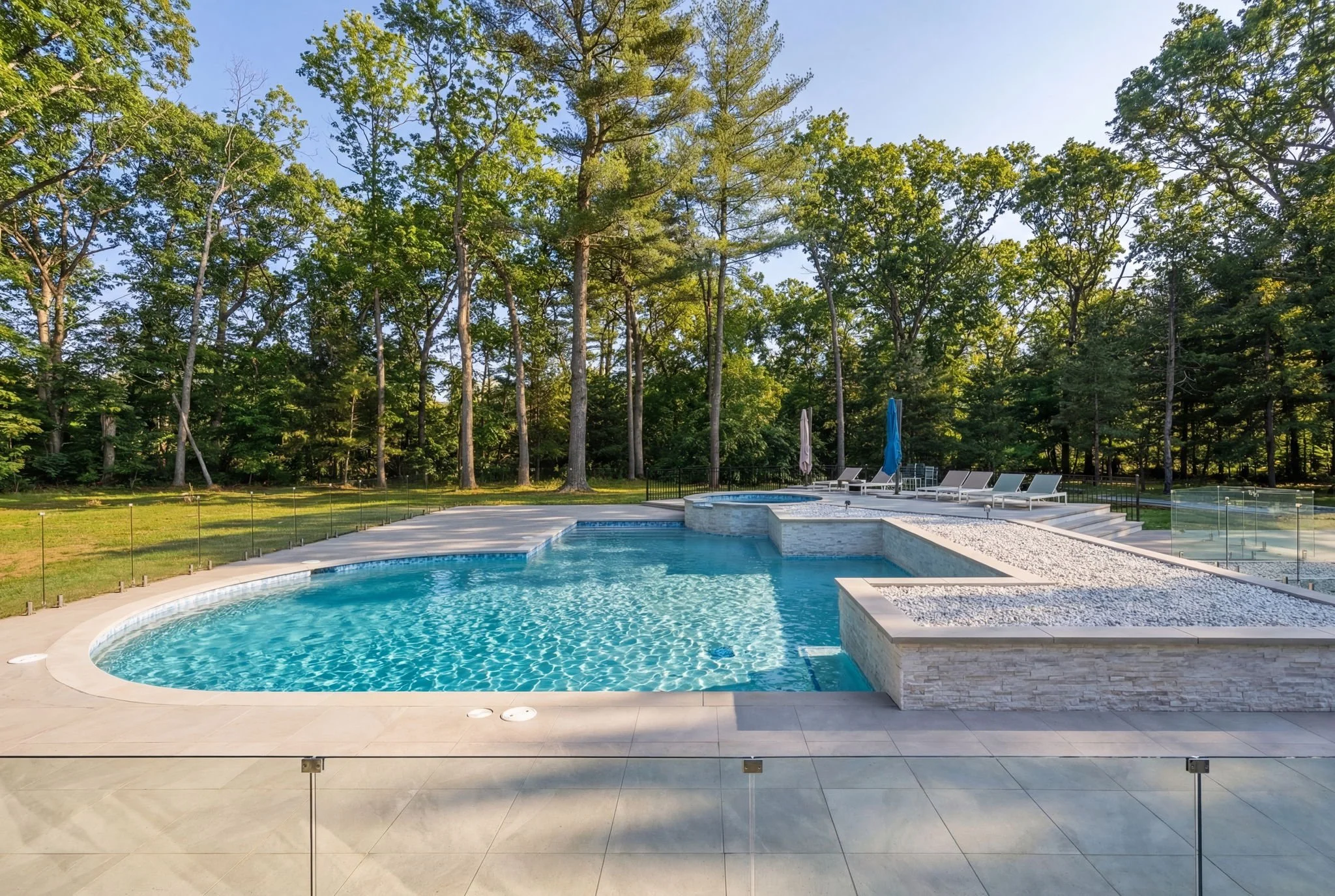 Swimming pool with surrounding lounge chairs and umbrellas, fenced with glass panels, set in a grassy area with tall trees and a clear sky in the background.