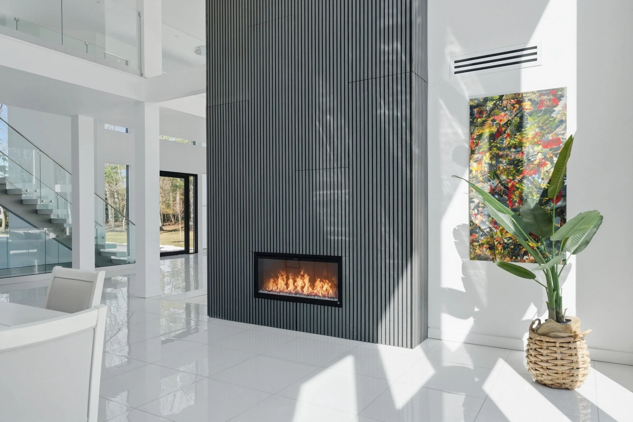 Modern living room with a black vertical panel fireplace, white tiled floor, white walls, a large colorful abstract painting, and a potted plant with large green leaves in a woven basket.