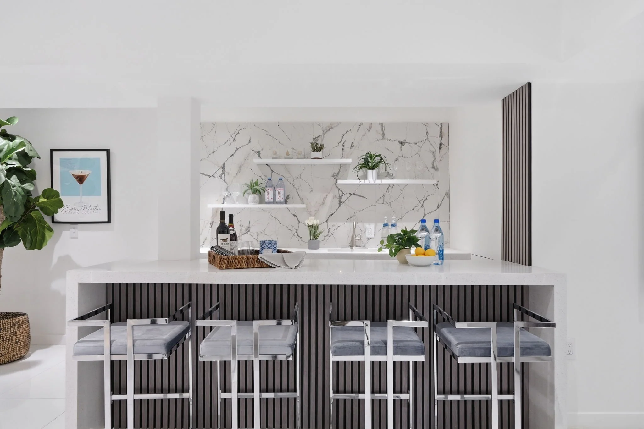 Modern kitchen with white marble backsplash, black and white striped bar, four metal and gray cushioned bar stools, decorative plants, bottles, and a bowl of lemons on the counter.