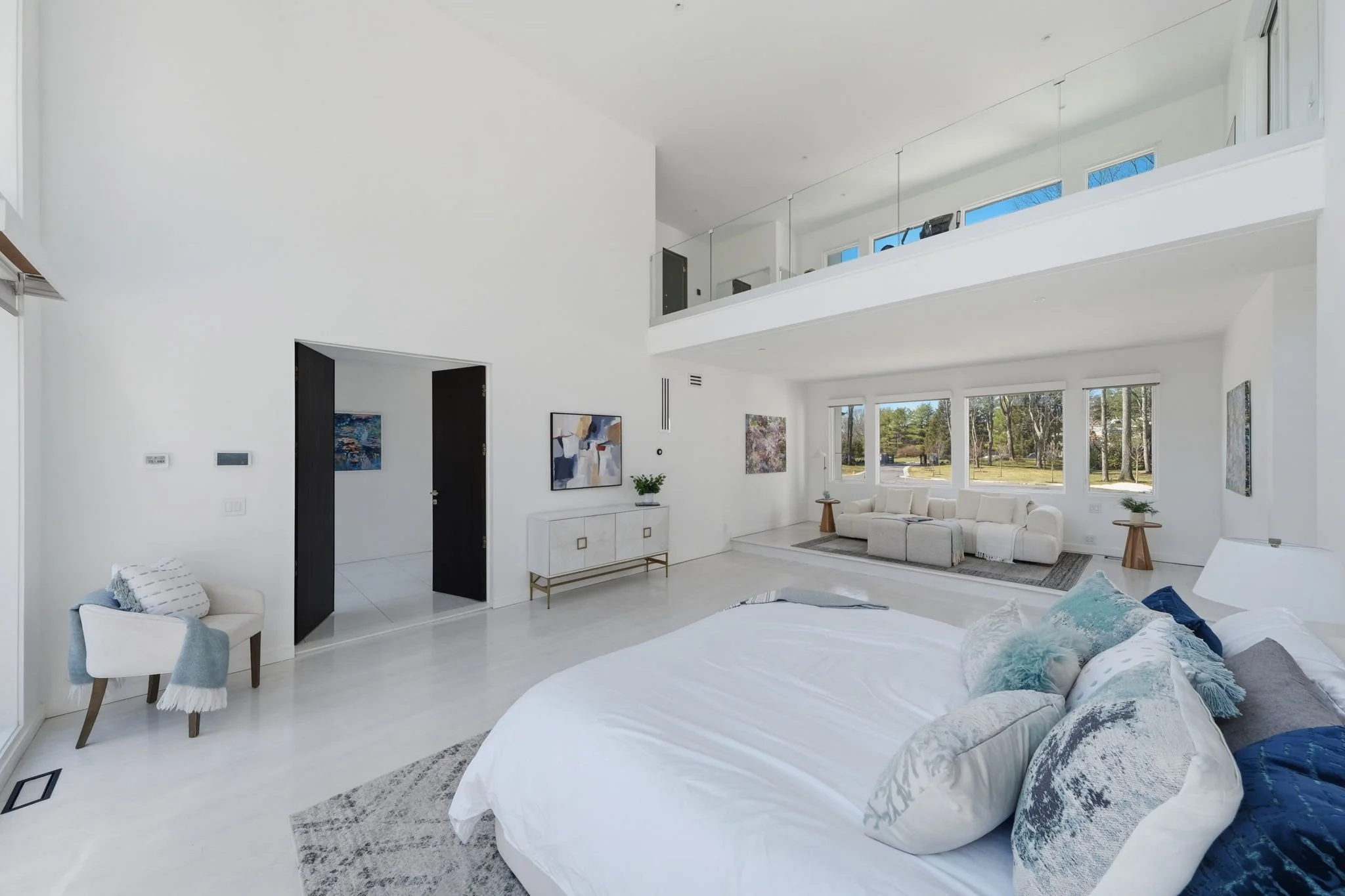 Bright, modern living room with white walls and furniture, large windows with a view of trees, and minimal decor including paintings and plants.