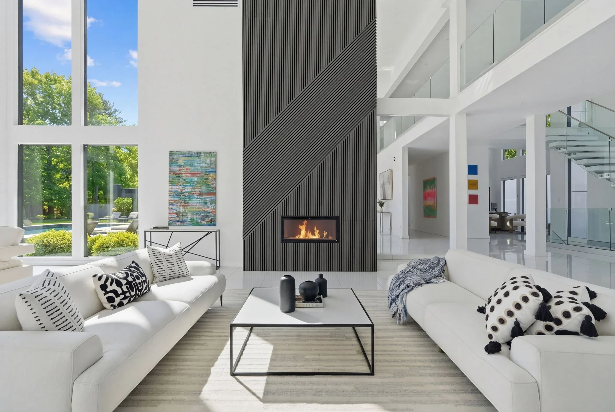Modern living room with two white sofas, black and white decorative pillows, a black coffee table with vases, a black textured fireplace, large windows showing greenery outside, and abstract artwork on the walls.