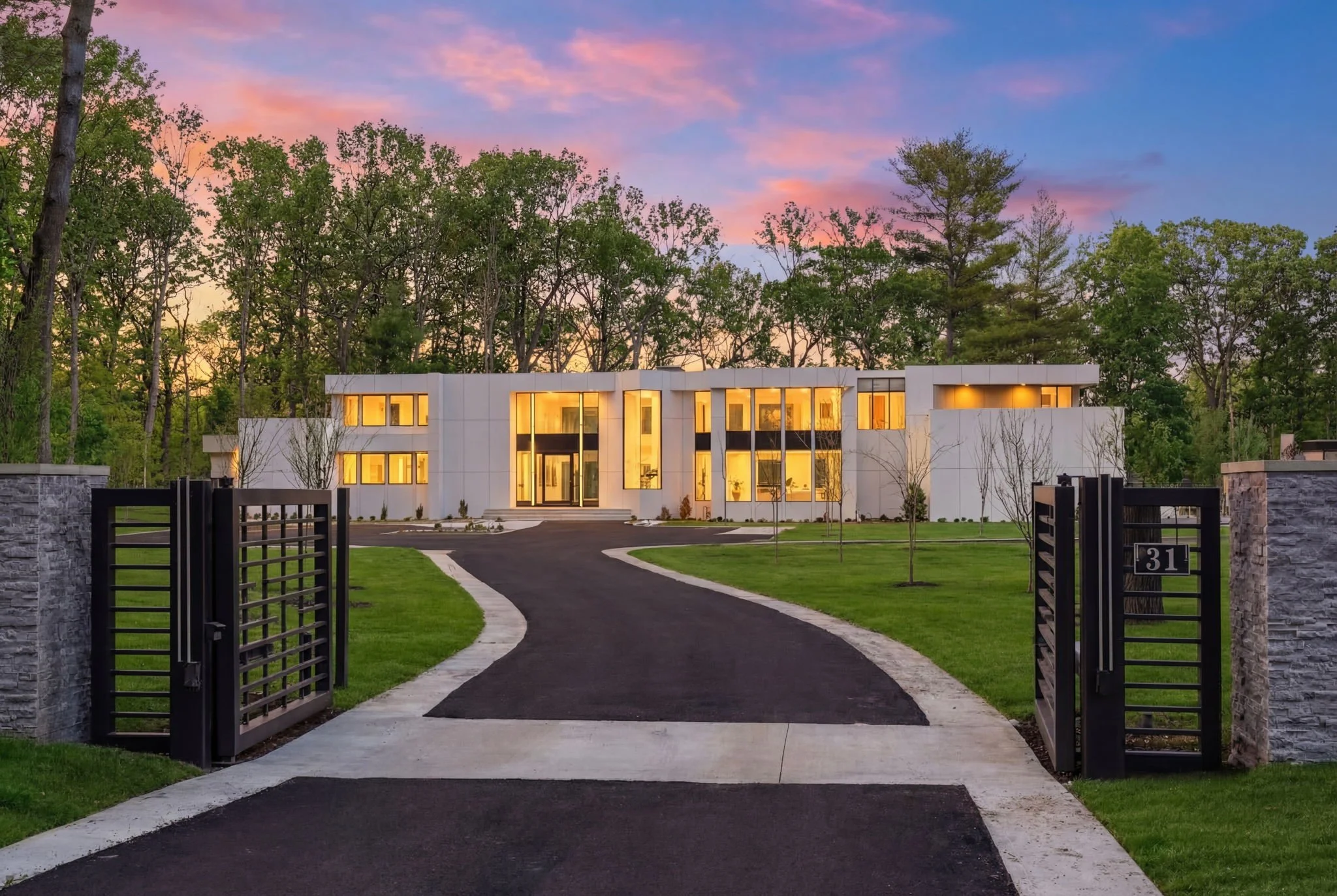 Modern white house with large glass windows, illuminated from inside, situated at the end of a winding asphalt driveway, surrounded by a green lawn and tall trees, during sunset.