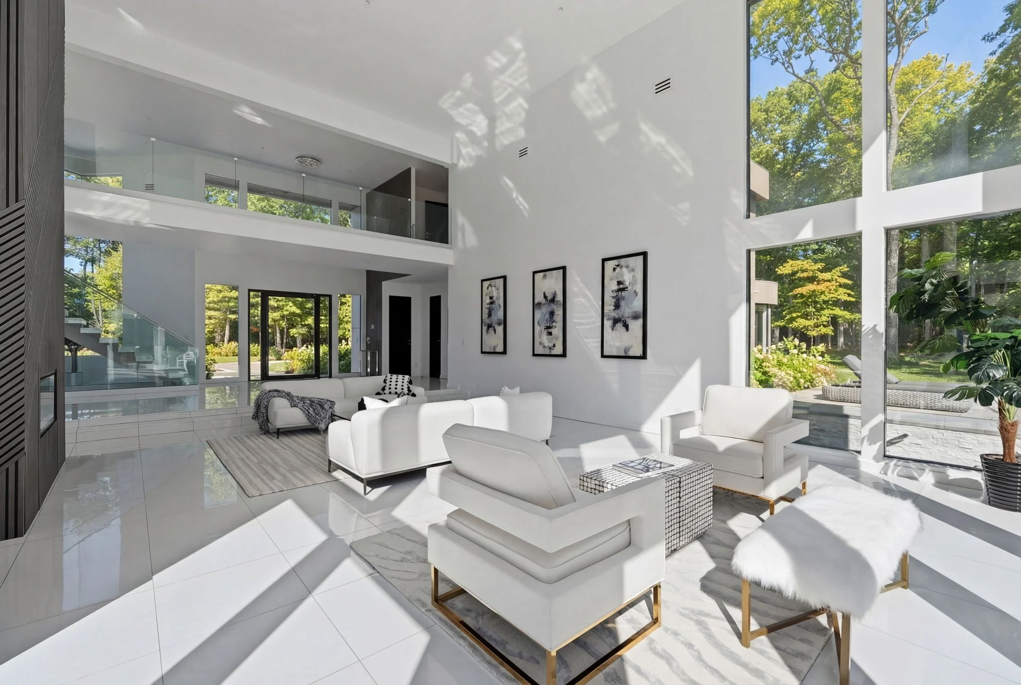 Modern living room with white furniture, large windows, and artistic wall decor, overlooking a lush green outdoor area.