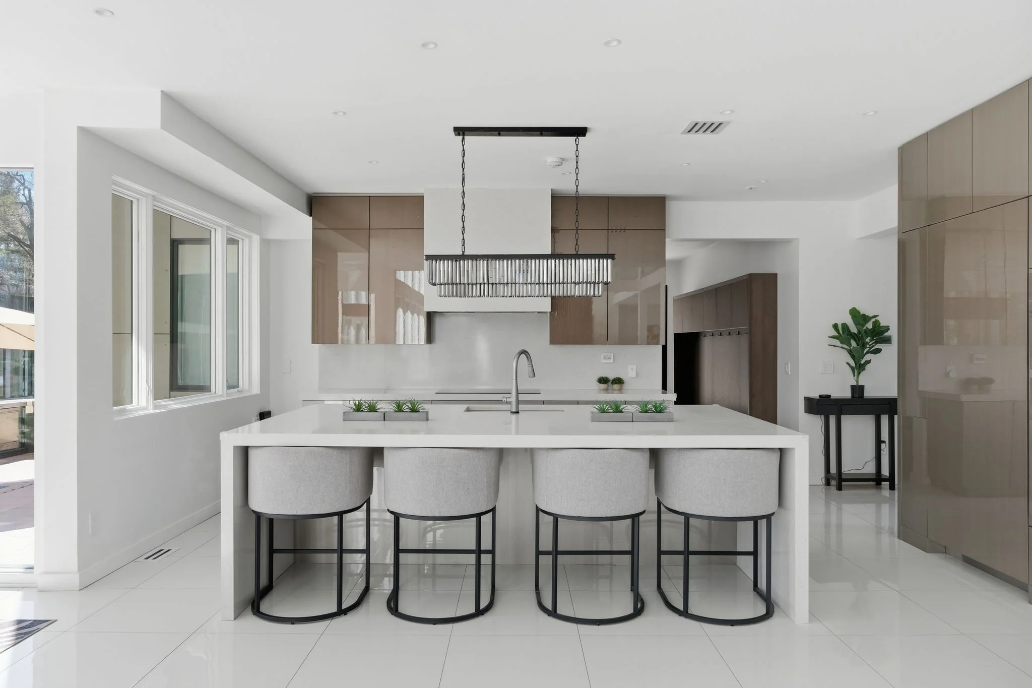 Modern kitchen with white countertops, four barstools, beige cabinets, and a black table with a plant.