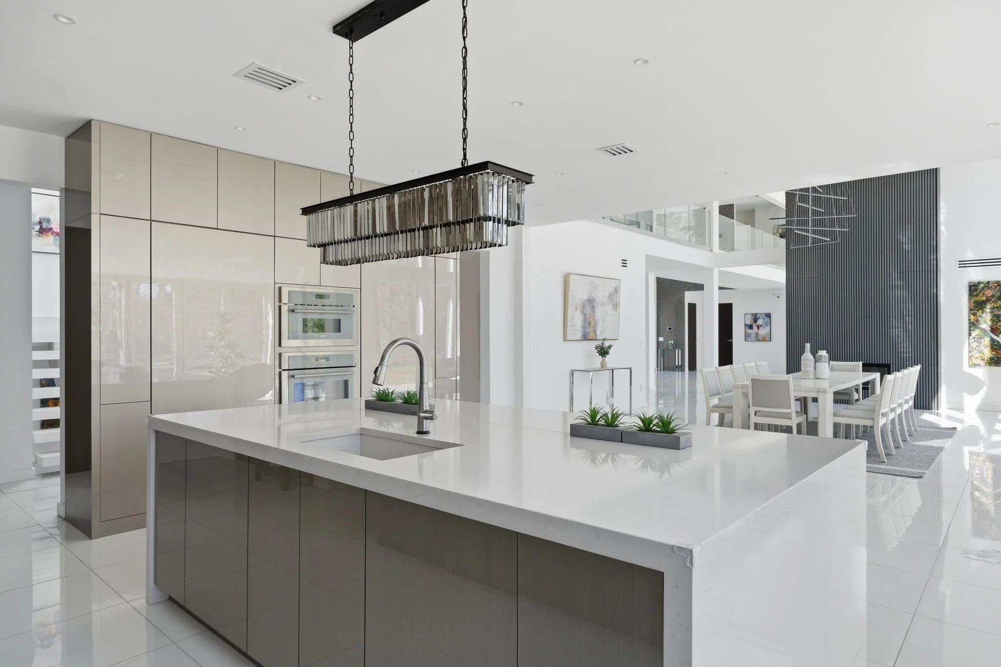 Modern open-concept kitchen and dining area with white walls, a central island with a sink, and a chandelier overhead. The dining space features a long table with white chairs, and artwork is displayed on the walls.