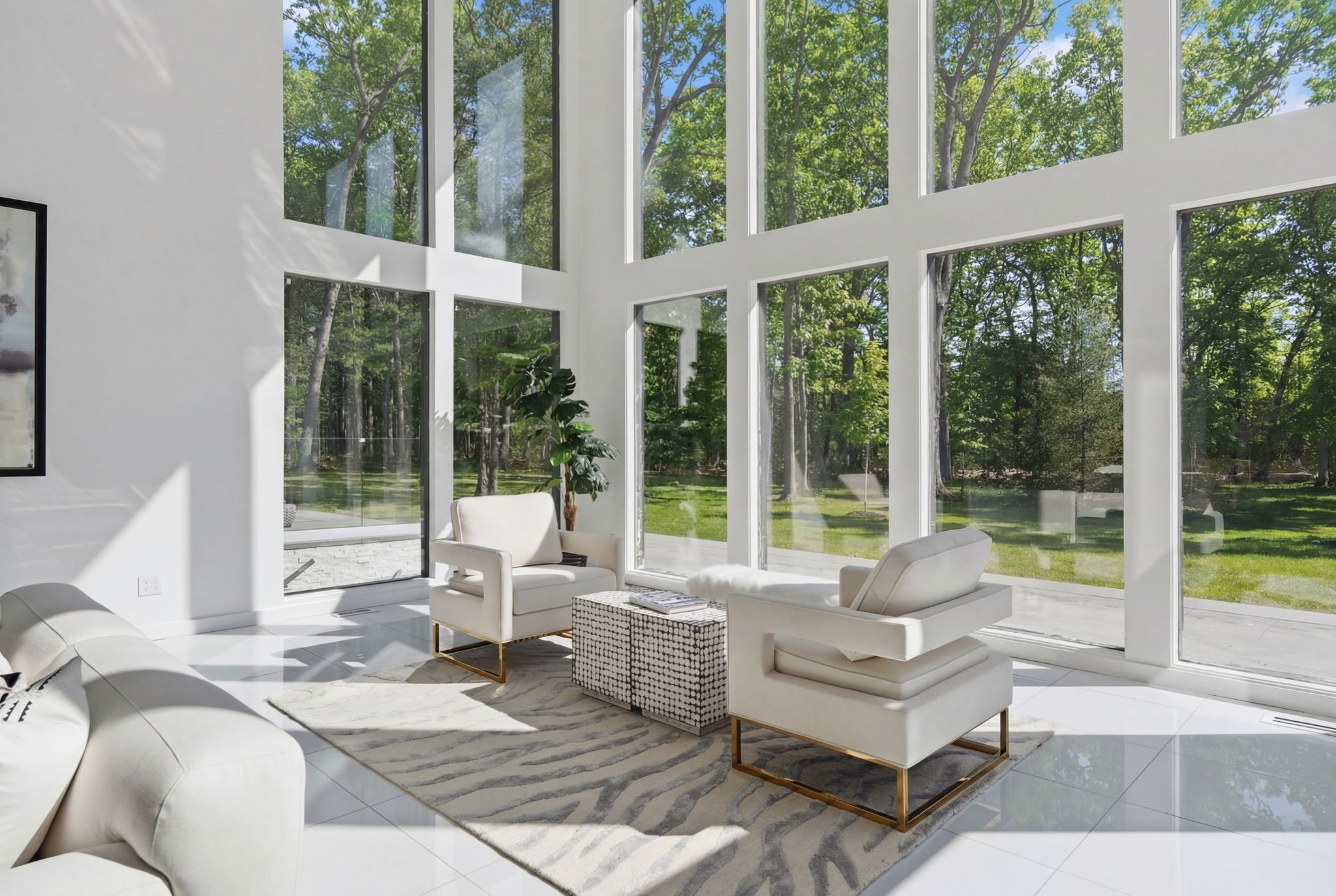 Bright living room with floor-to-ceiling windows overlooking a wooded yard, featuring white sofas, a plant, a patterned ottoman, and a zebra-patterned rug.
