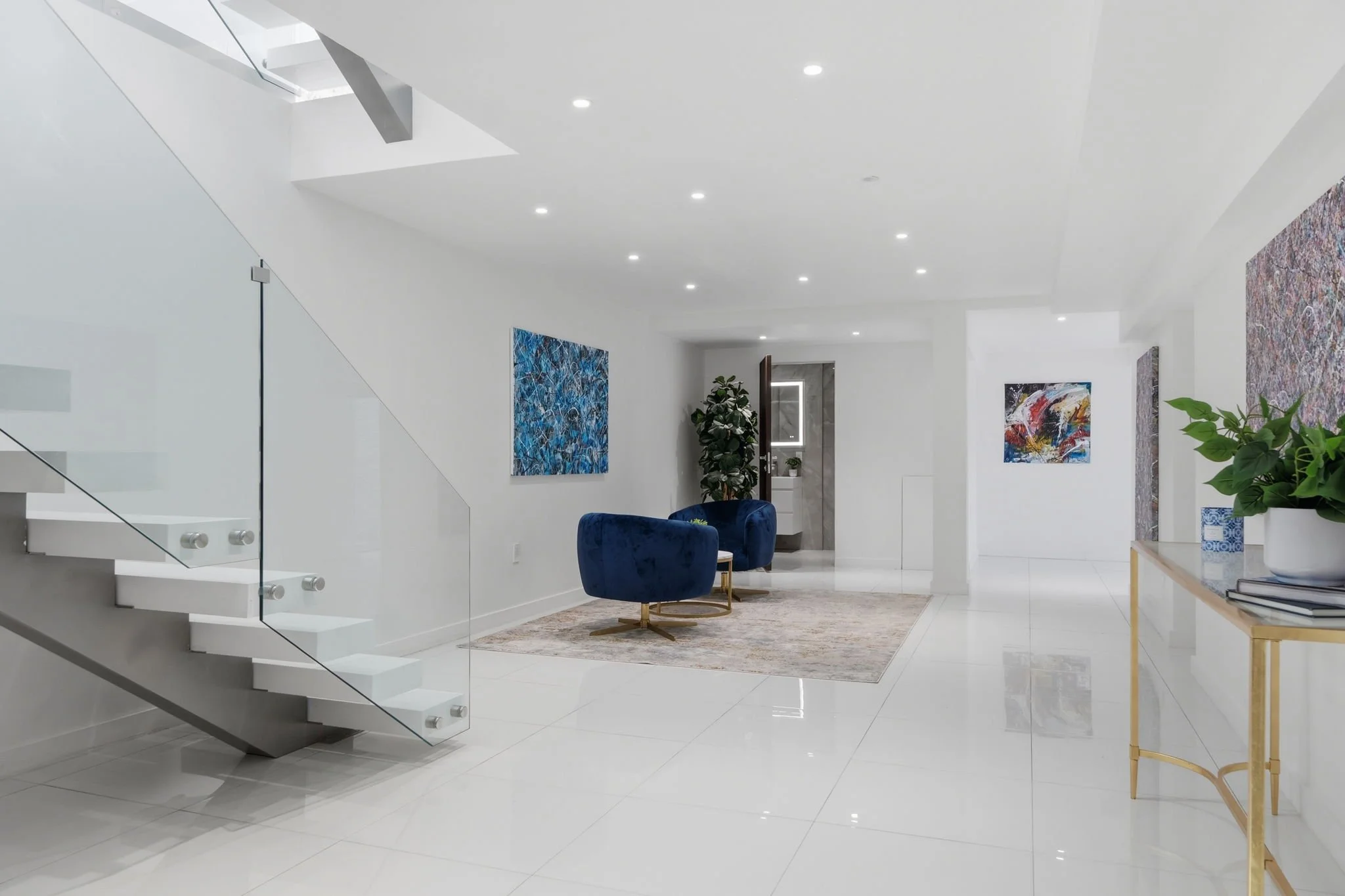 Modern, minimalist living room with white walls and glossy white tiles, blue velvet armchairs, colorful abstract art, a gold and glass side table, and a potted plant near a doorway.