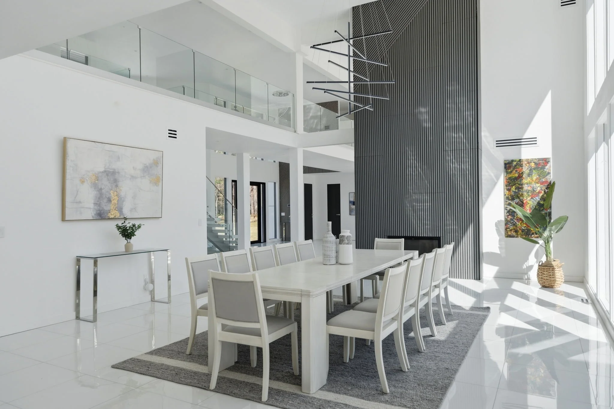 Modern dining room with a long white table, white chairs, a gray rug underneath, and contemporary decor including vases and plants. Large windows let in natural light, illuminating the space.