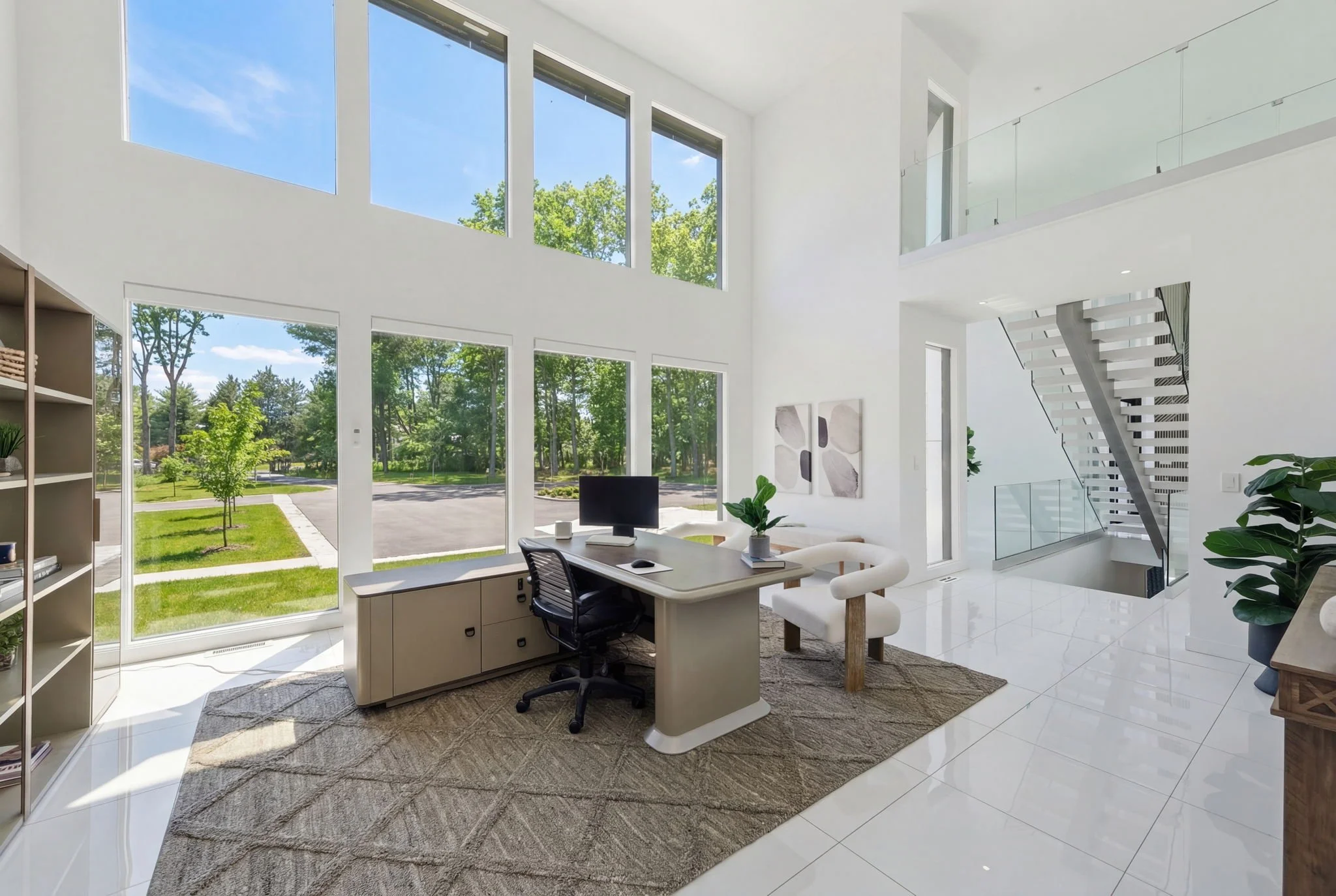 Bright modern office with large floor-to-ceiling windows overlooking a lush green yard, white walls, a desk with a computer and a black office chair, a beige rug, and decorative artwork on the wall.
