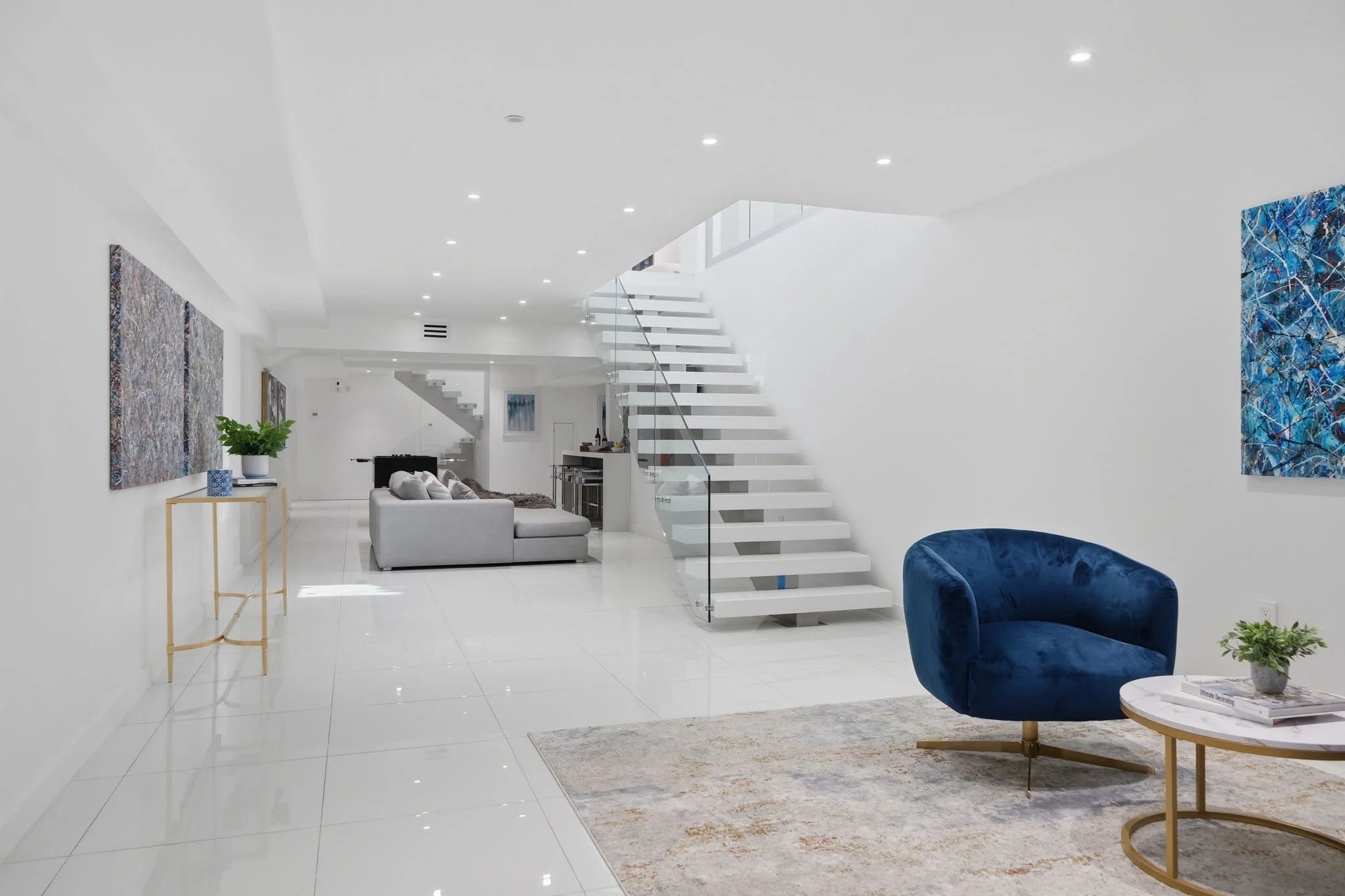 Modern, minimalist living room with white walls and glossy white tile flooring. Includes a blue velvet armchair, a white and gold coffee table, a grey sofa, colorful abstract wall art, and a staircase with glass railing leading upstairs.