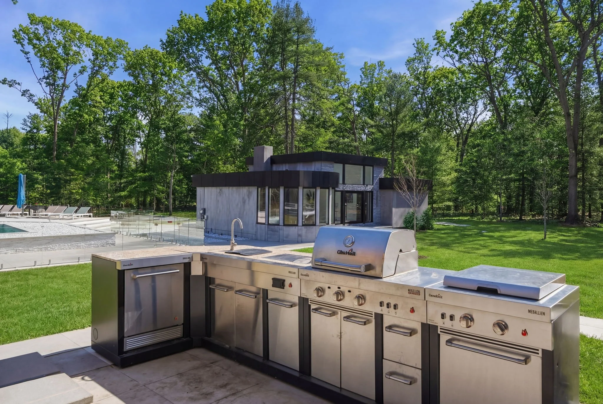 Outdoor kitchen with stainless steel grill, sink, and refrigerator in a backyard with a lawn, trees, and a modern house in the background.