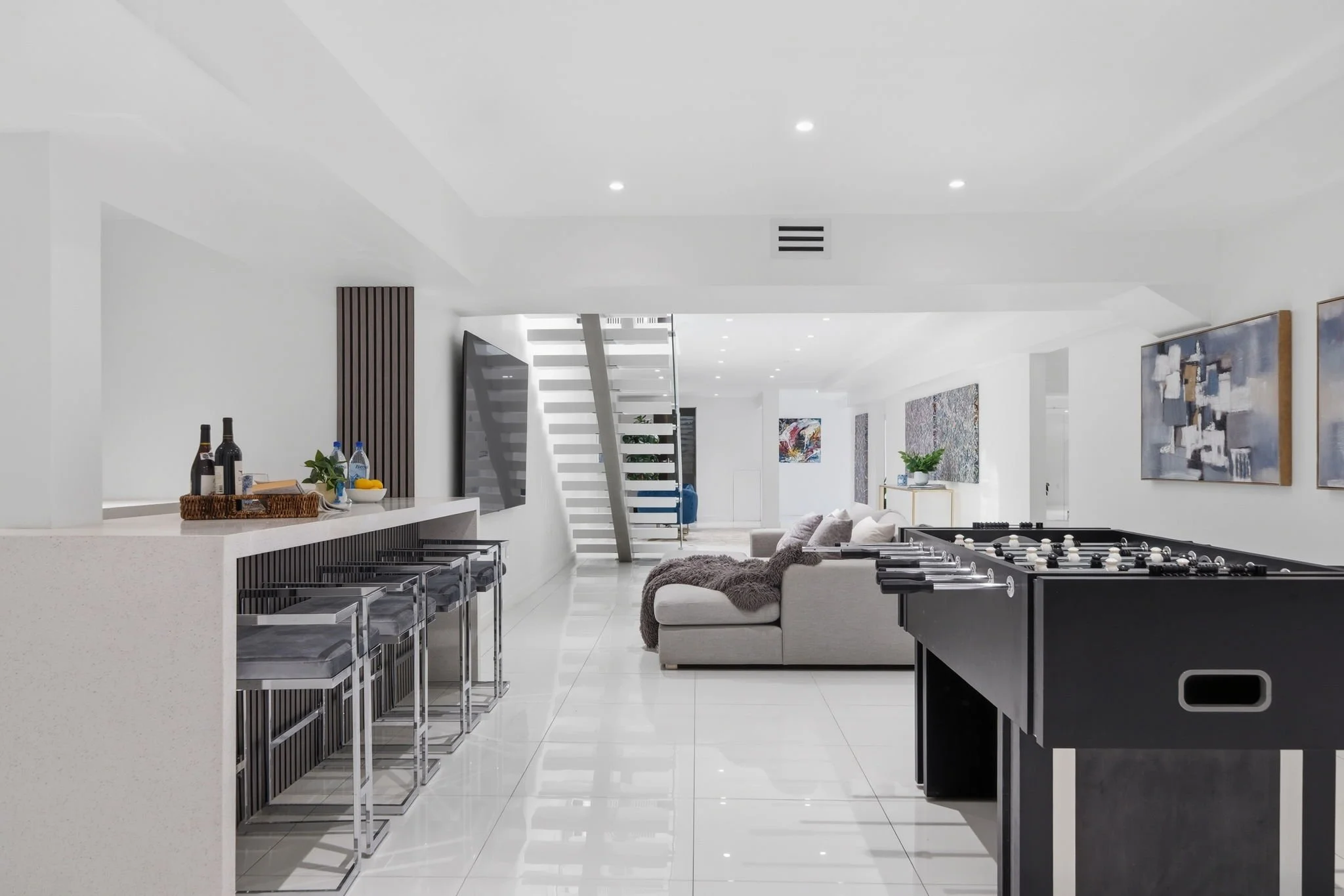 Modern living room with white walls, tiled floor, and contemporary furniture, including a gray sectional sofa with a faux fur throw, abstract paintings on the walls, and a foosball table.