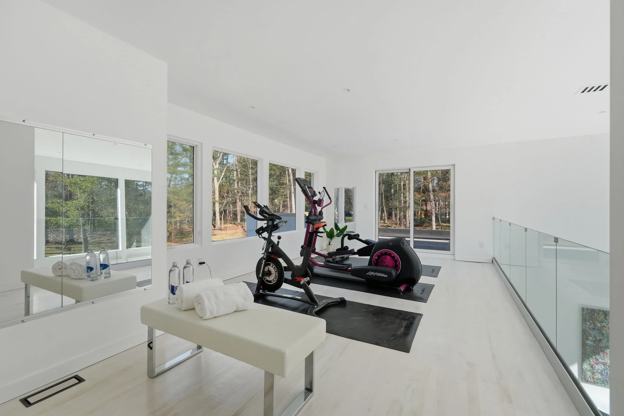 Modern home gym with two exercise machines and large windows overlooking trees.