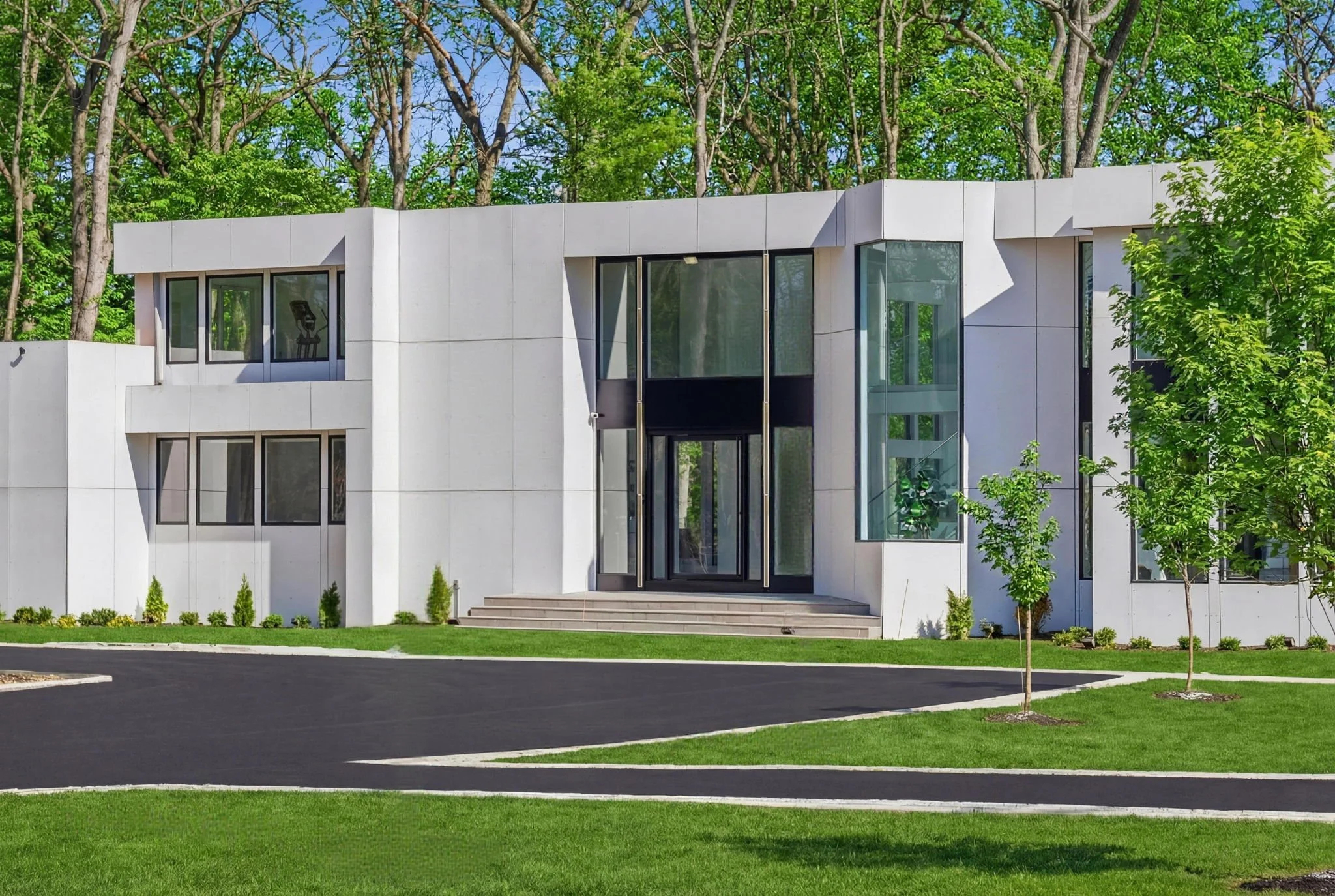 Modern white house with large glass windows and a landscaped yard with green grass and small trees.